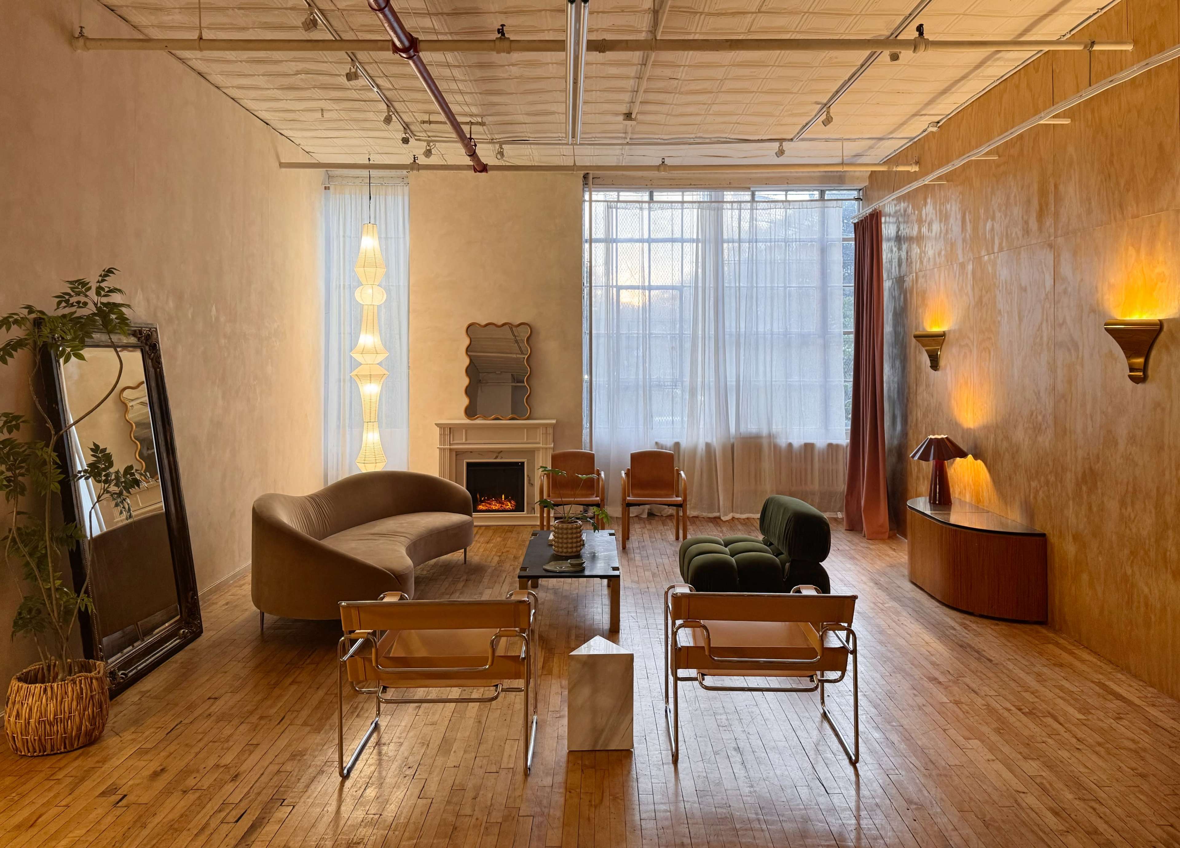 The image shows a spacious, modern living area featuring a curved sofa, two armchairs, a coffee table, a reflective mirror, and various light fixtures along the walls.