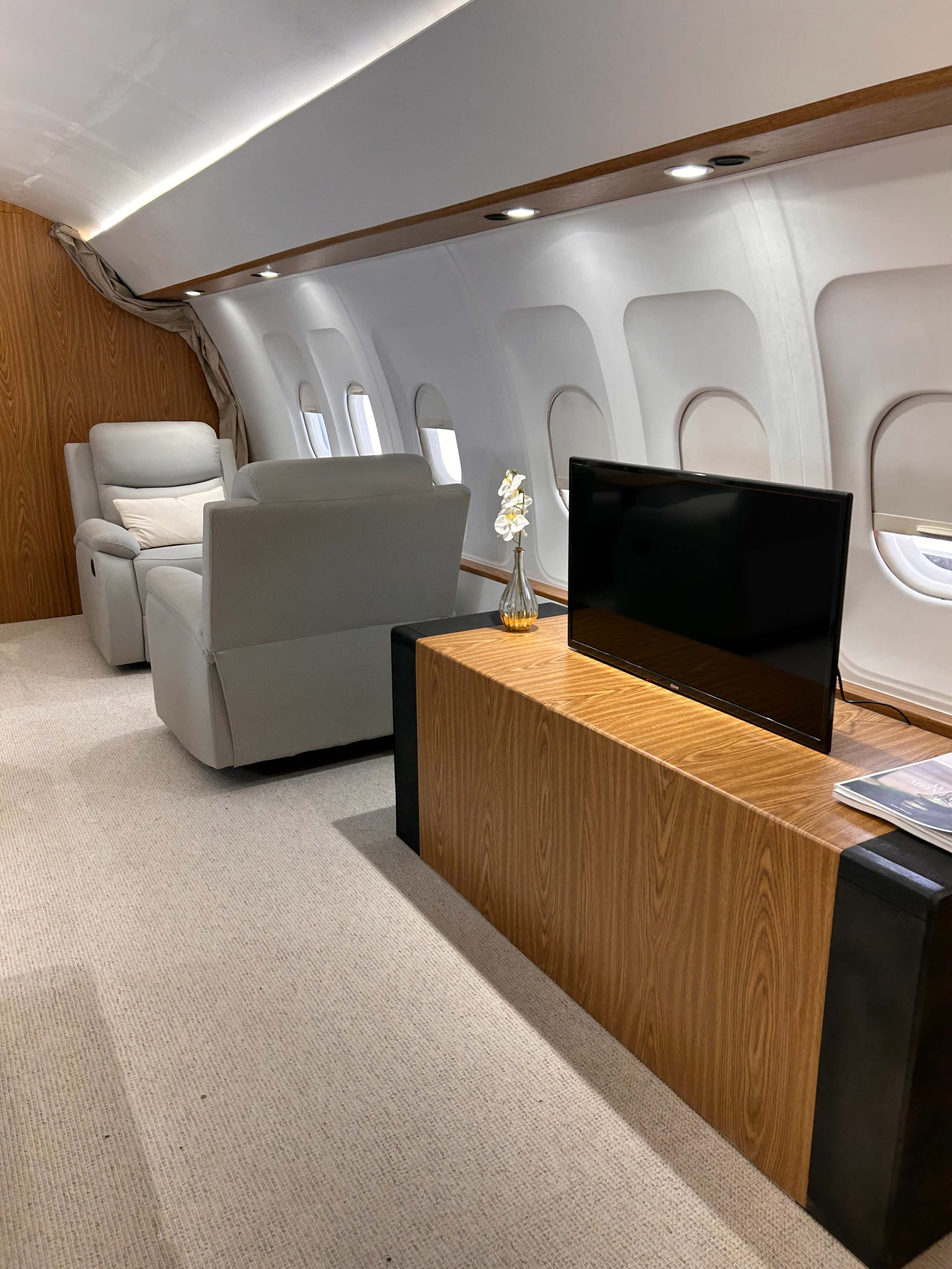 A luxurious interior of a private jet, featuring a reclining chair, a television on a wooden console, and large windows.