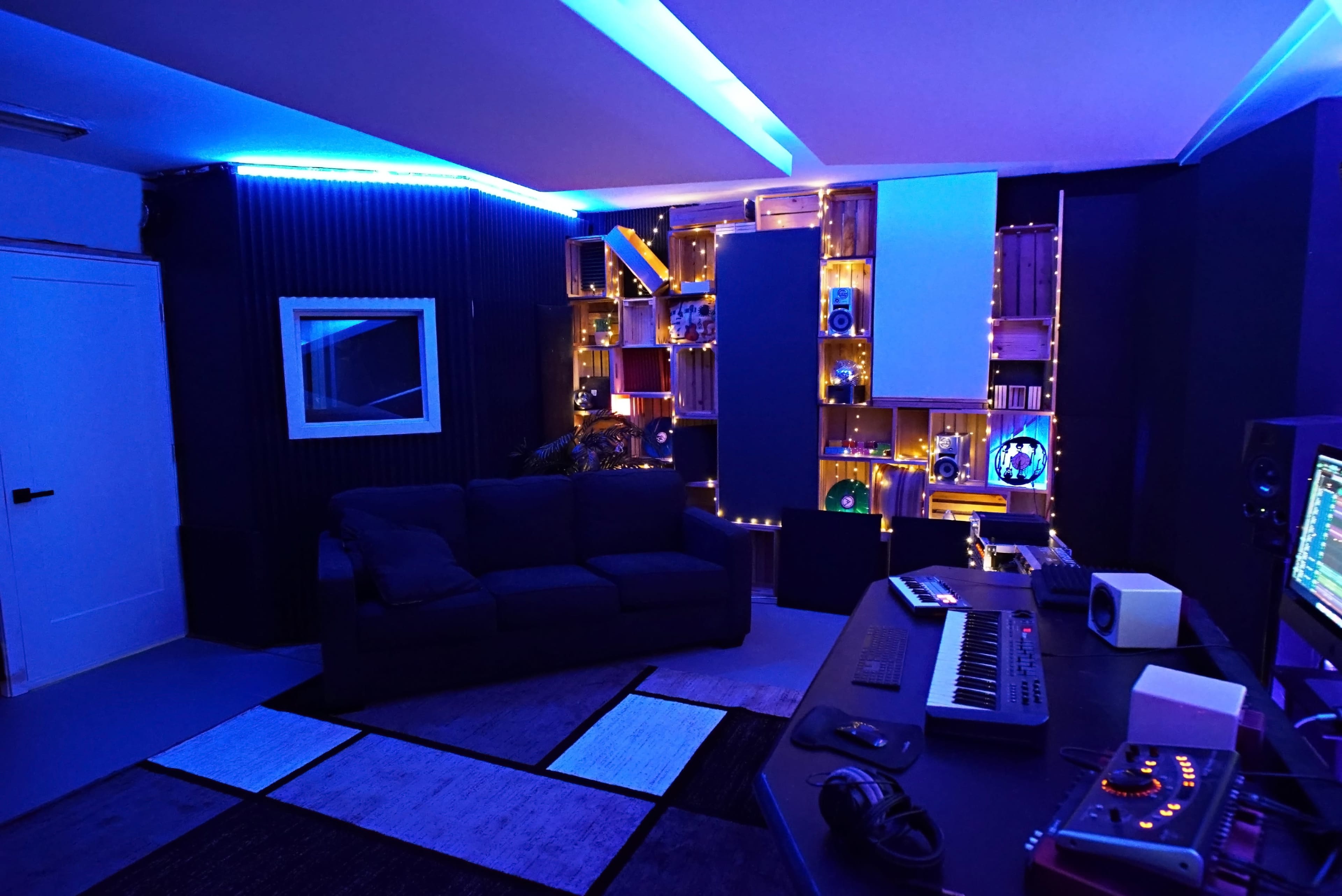 A music production studio features a dark blue color scheme with ambient lighting, a couch, and electronic equipment on a desk.