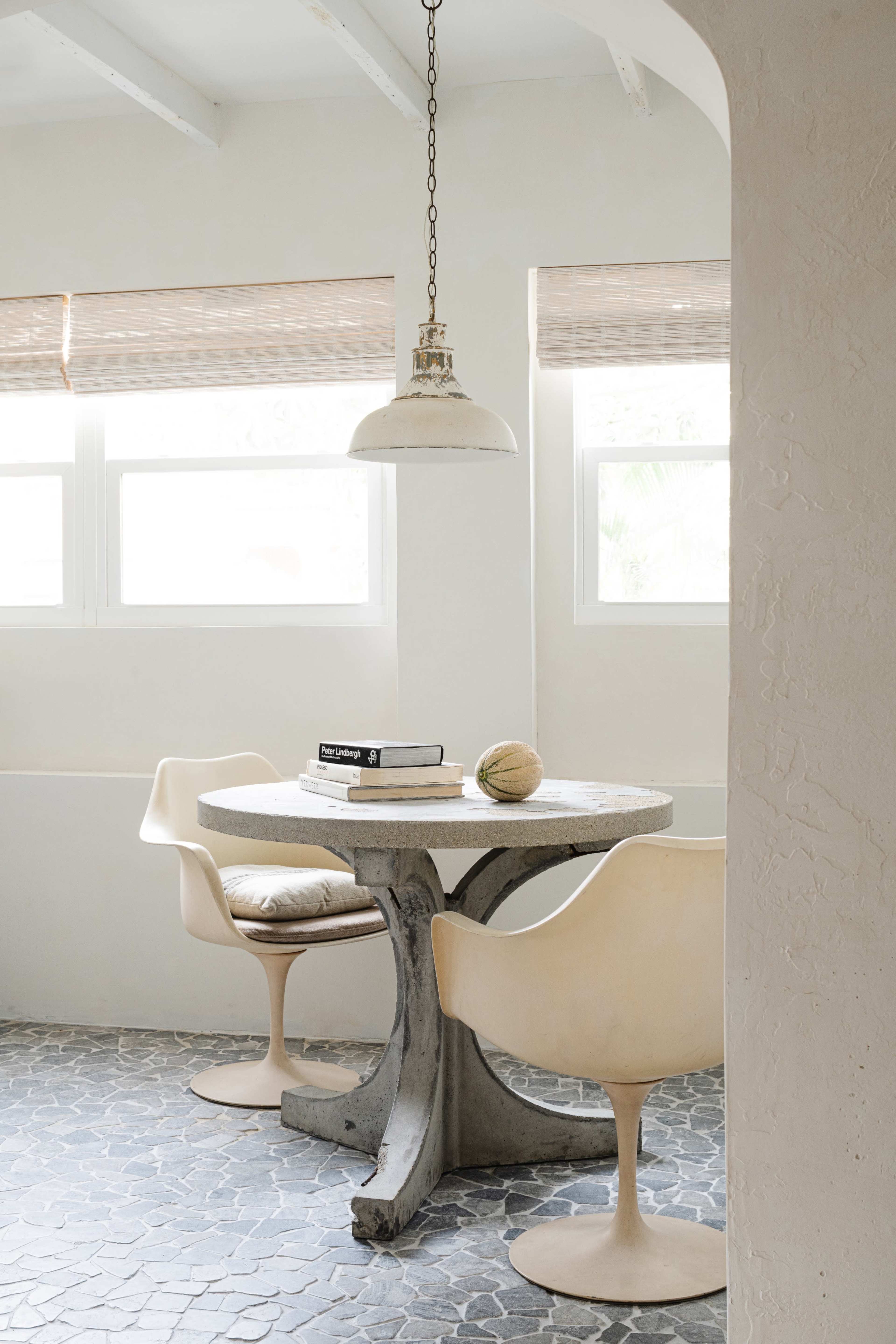 A round table with two chairs is positioned near a window, illuminated by a hanging light fixture, with books on the table and a textured floor beneath.