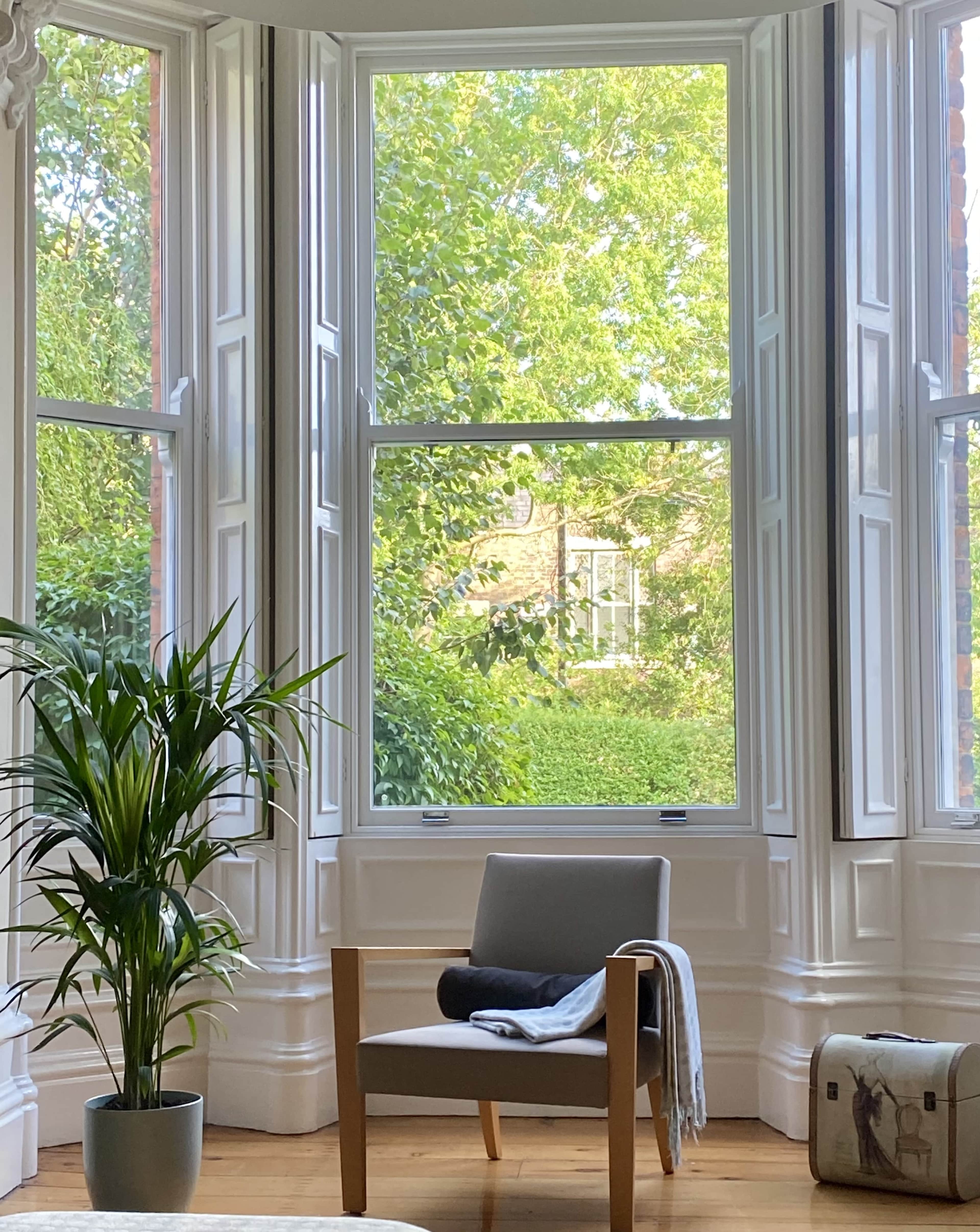 A cozy corner features a chair with a blanket and a potted plant in front of a large window overlooking greenery.