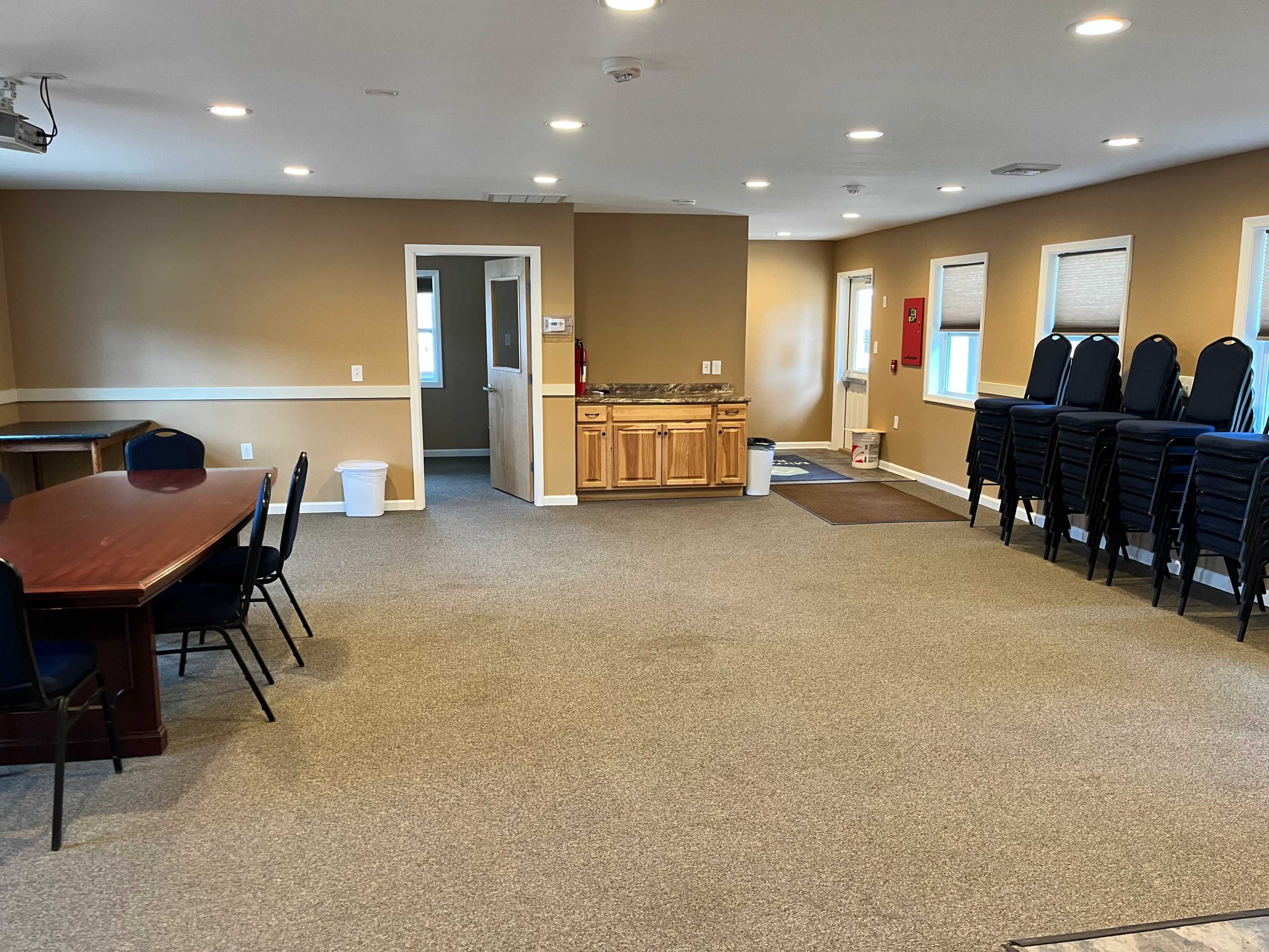 The image shows a spacious, empty room with a table and chairs on one side and a kitchenette area along the wall, while the opposite side features stacked chairs.