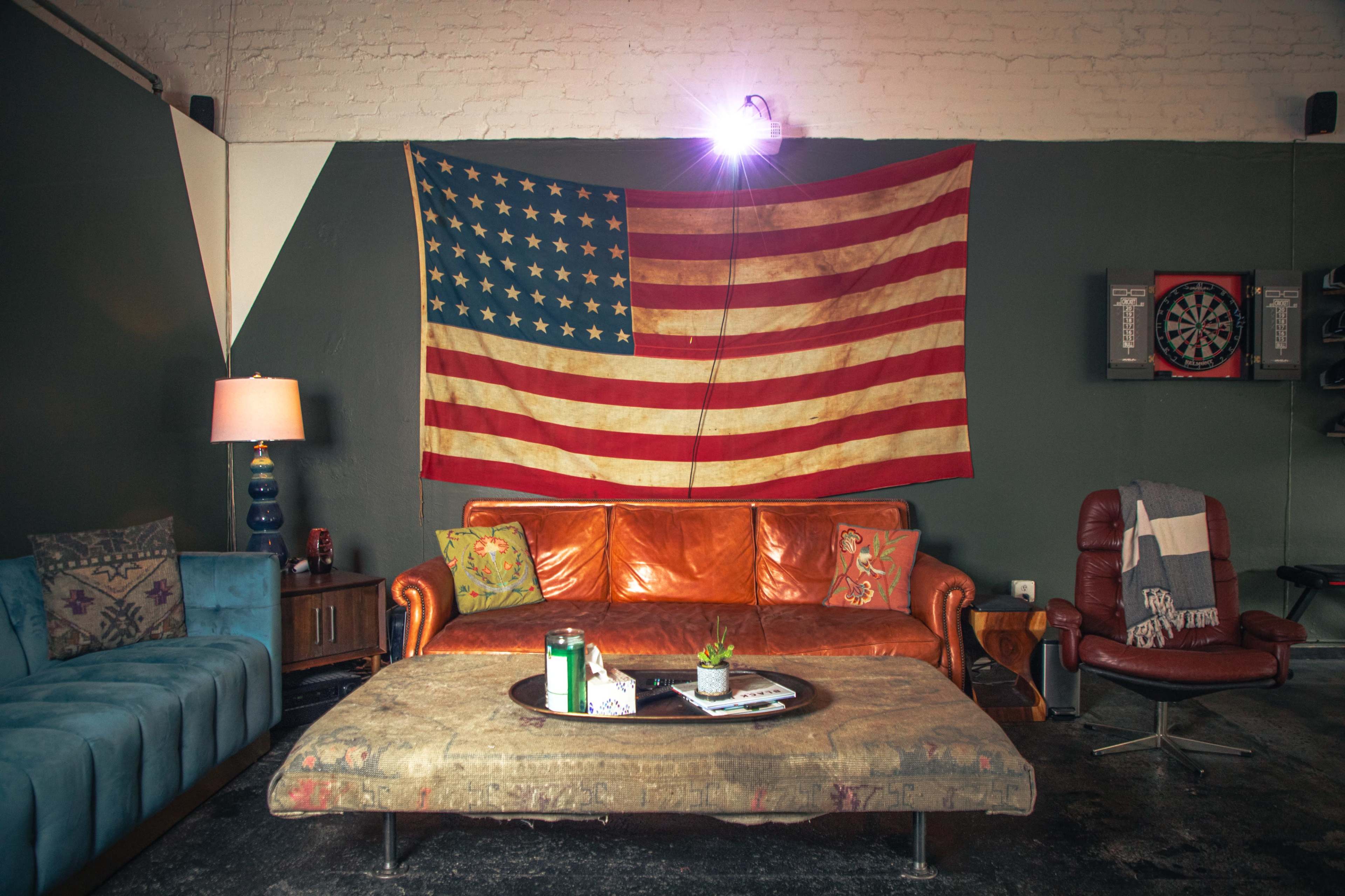 The image shows a cozy living room with a brown leather couch, a vintage American flag hanging on the wall, and a coffee table with a small plant.