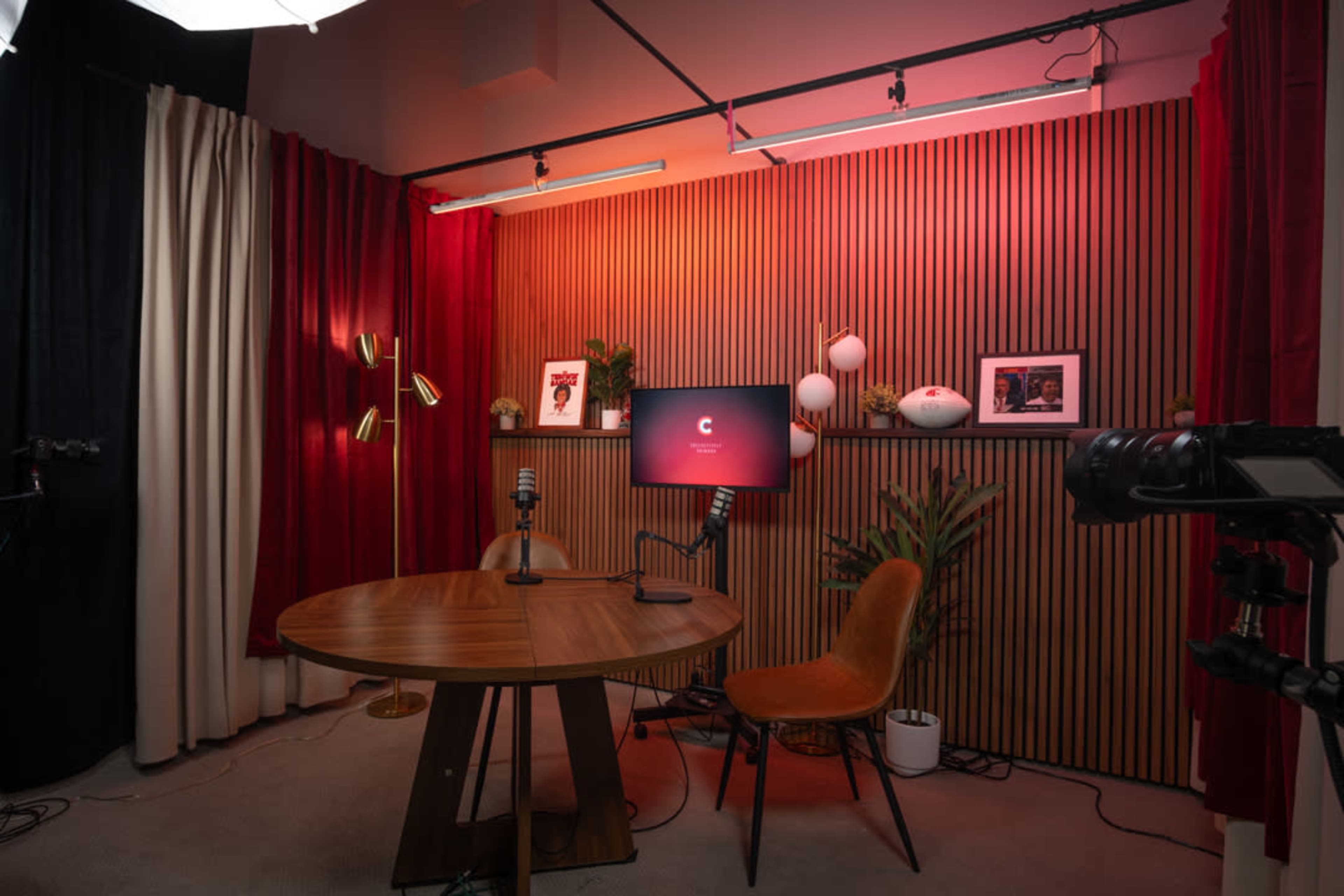 The image shows a modern studio setup featuring a round wooden table, a single orange chair, two microphones, and a television screen against a backdrop of red and black curtains with decorative elements.