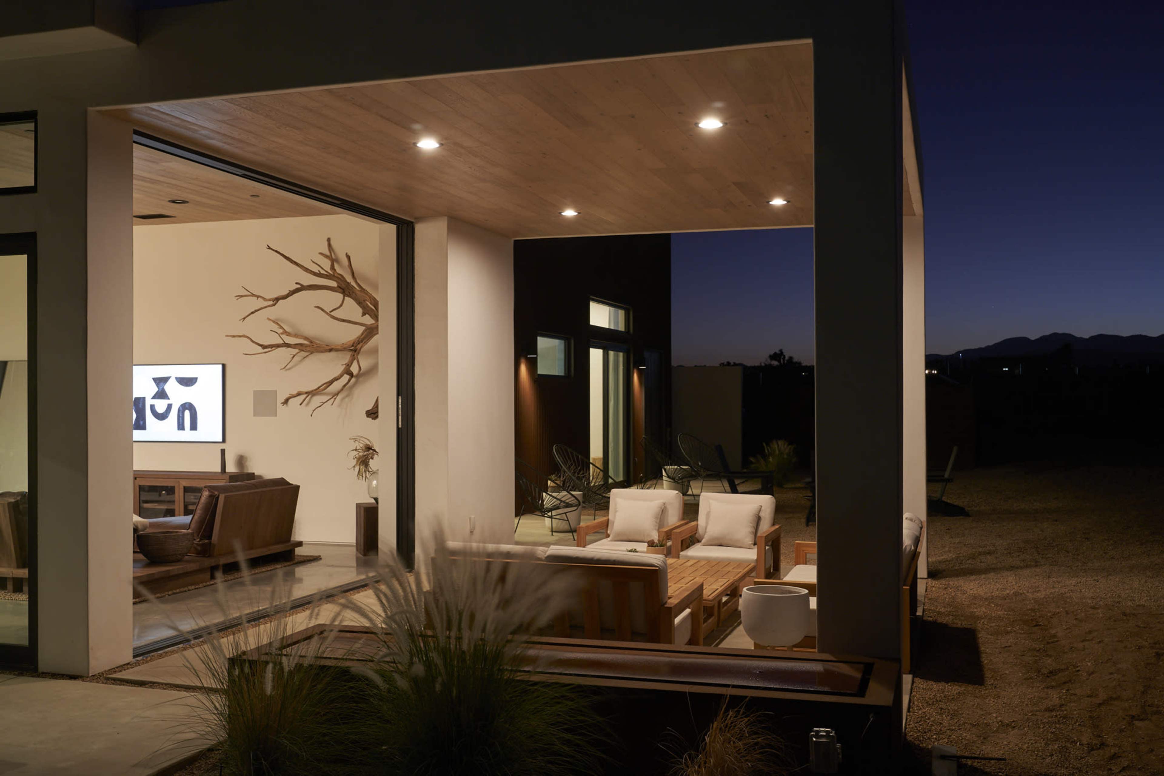The image depicts a modern outdoor living area with a minimalist design, featuring comfortable seating and a television visible through large glass doors, set against a twilight backdrop.