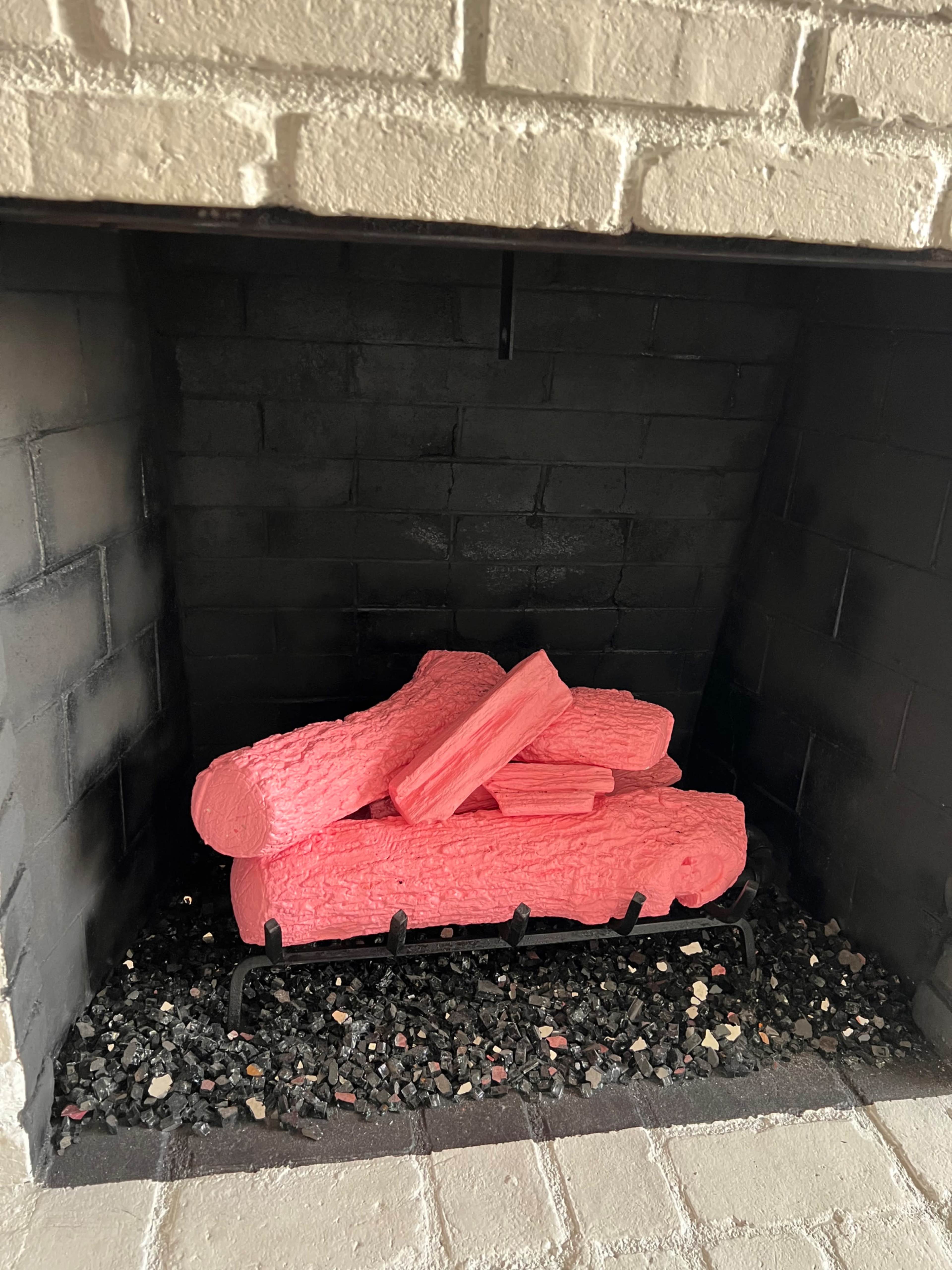 A pile of bright pink faux logs is arranged in a black fireplace with a bed of small colored stones.