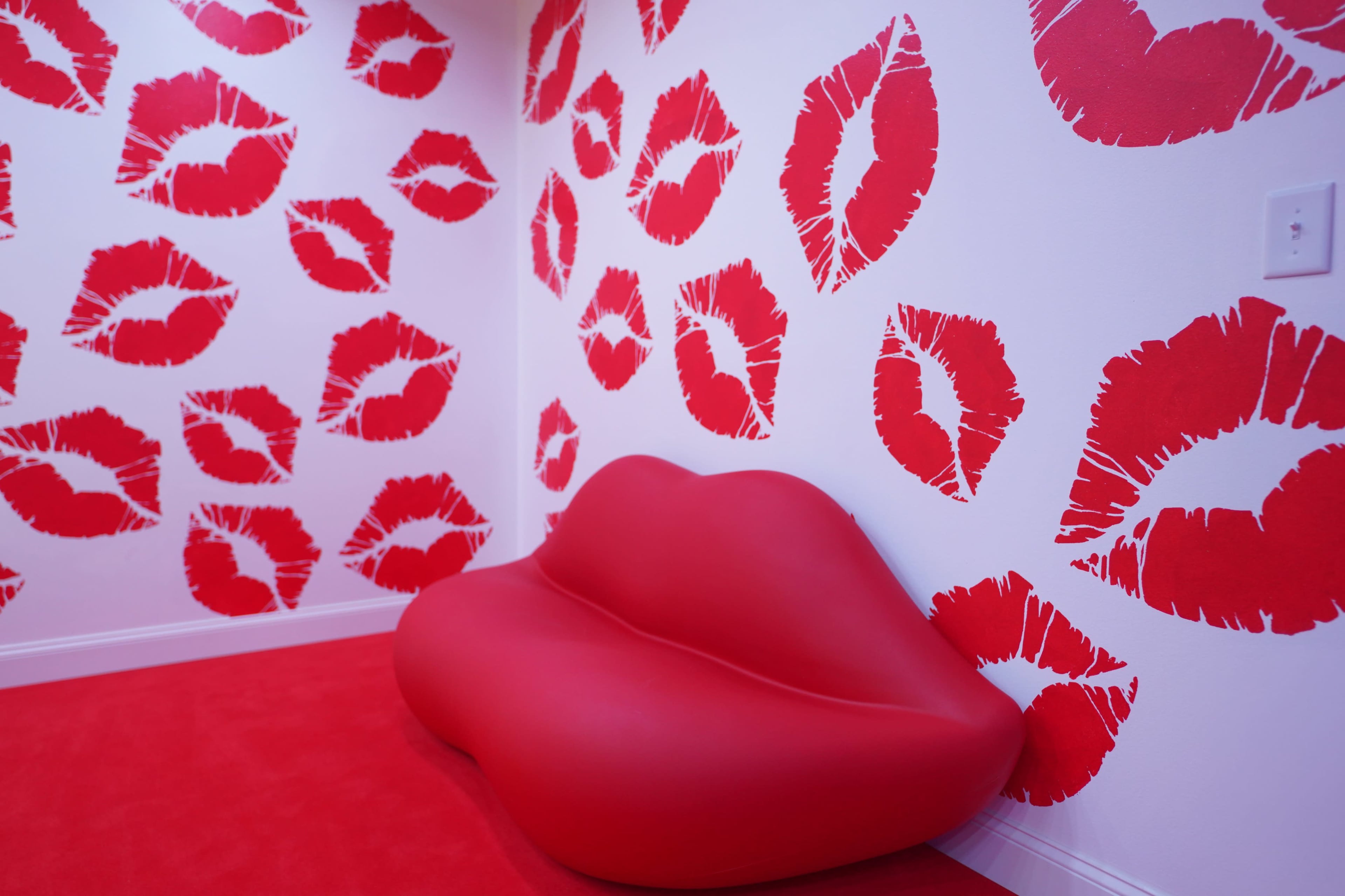 A room with red lipstick kiss prints on the white walls and a red lip-shaped sofa on a matching red carpet.
