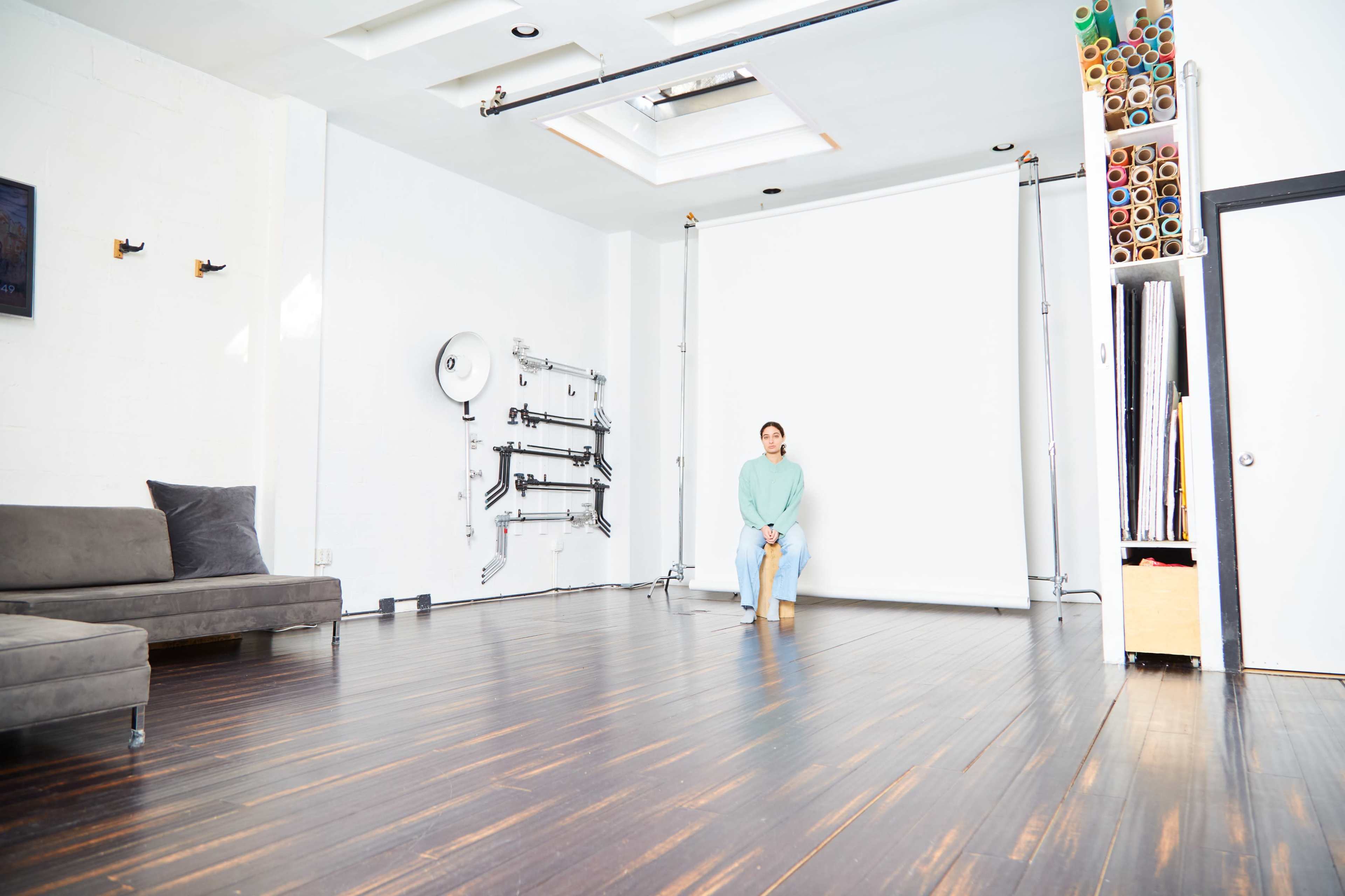 A person sits on a stool in a spacious, brightly lit photo studio with a white backdrop and wooden flooring.