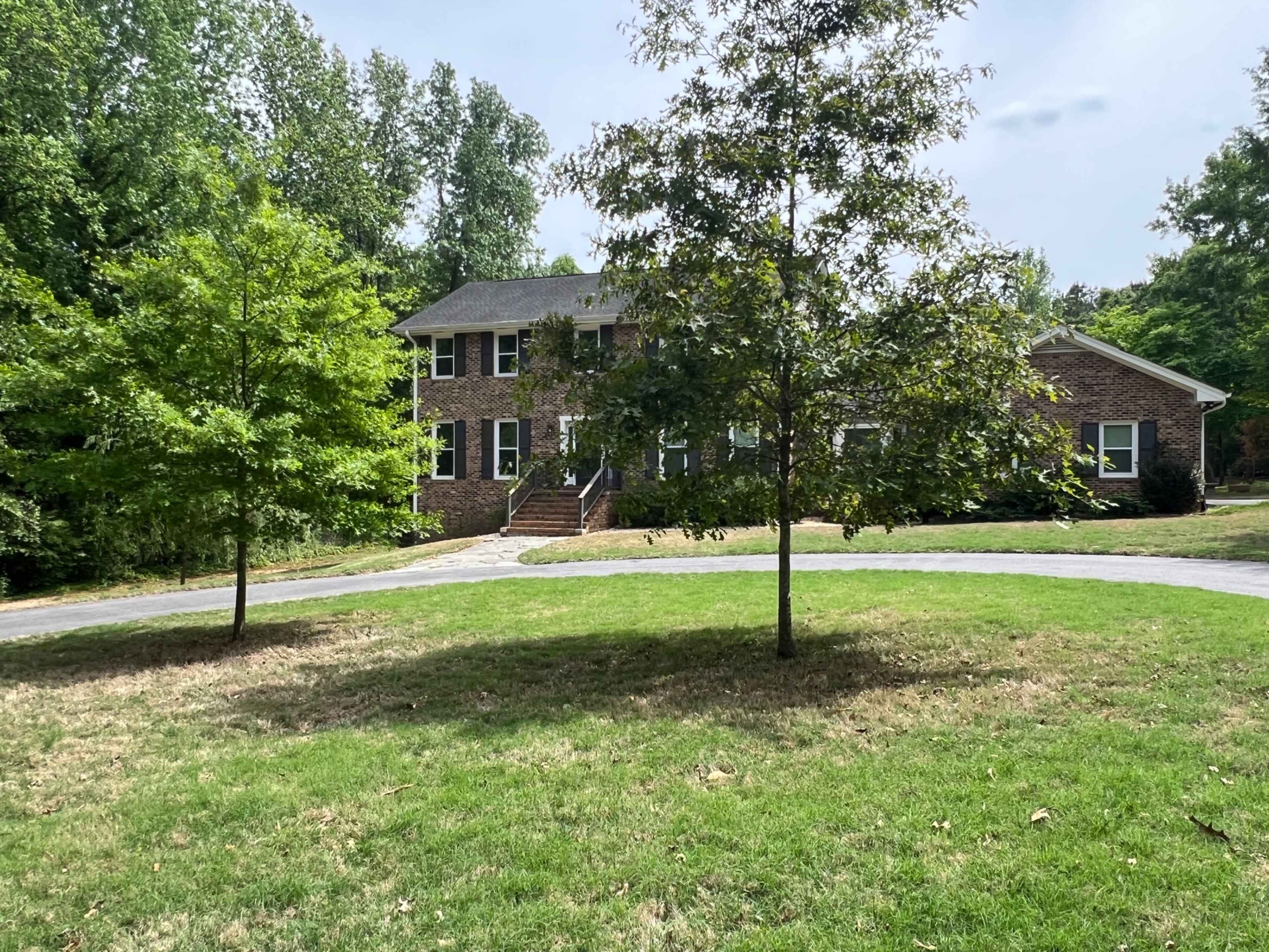 A two-story brick house is set back from a circular driveway, surrounded by mature trees and a well-maintained lawn.