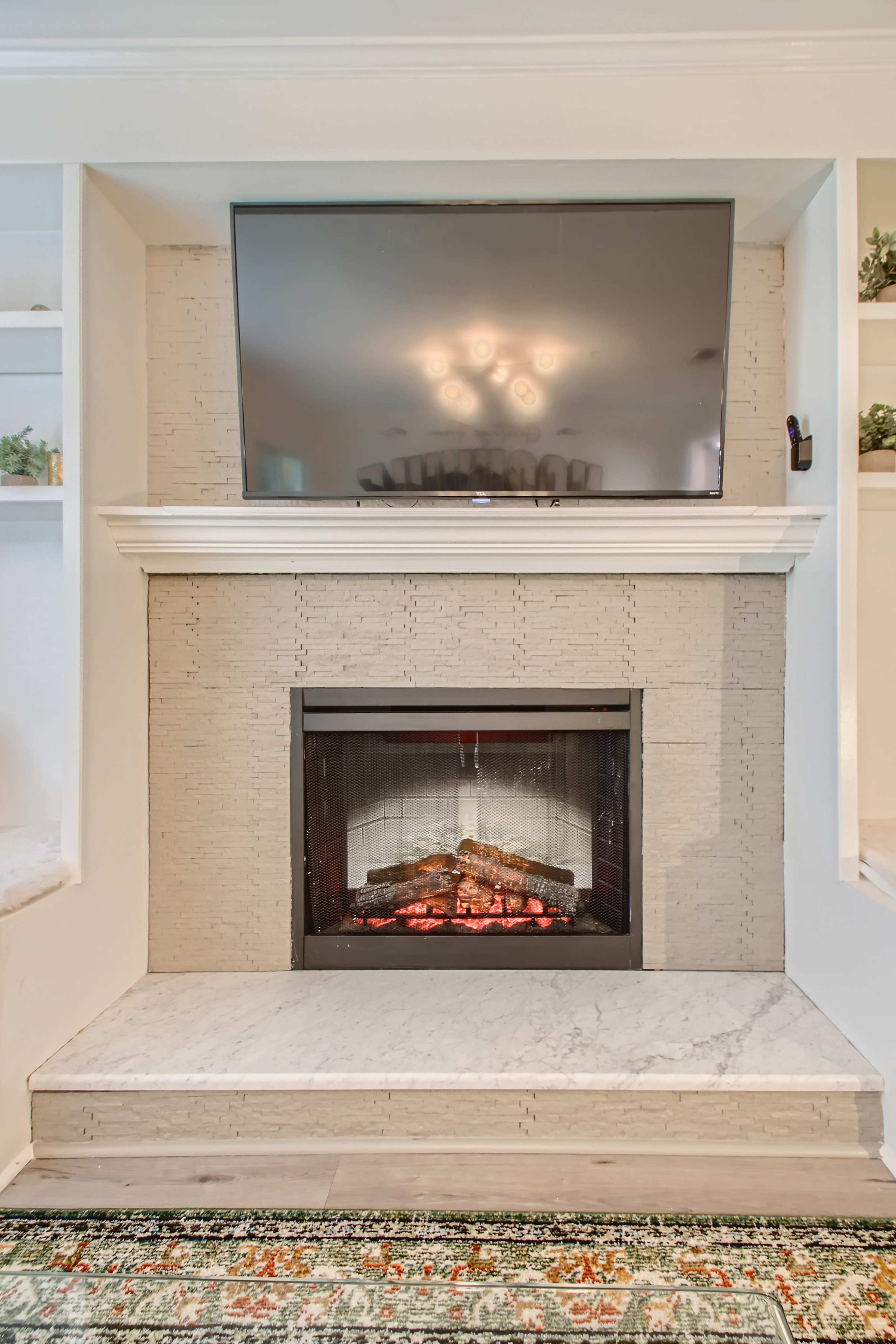 A modern fireplace with an electric insert sits beneath a wall-mounted television, framed by a light-colored stone surround and a marble base, against a backdrop of built-in shelves.