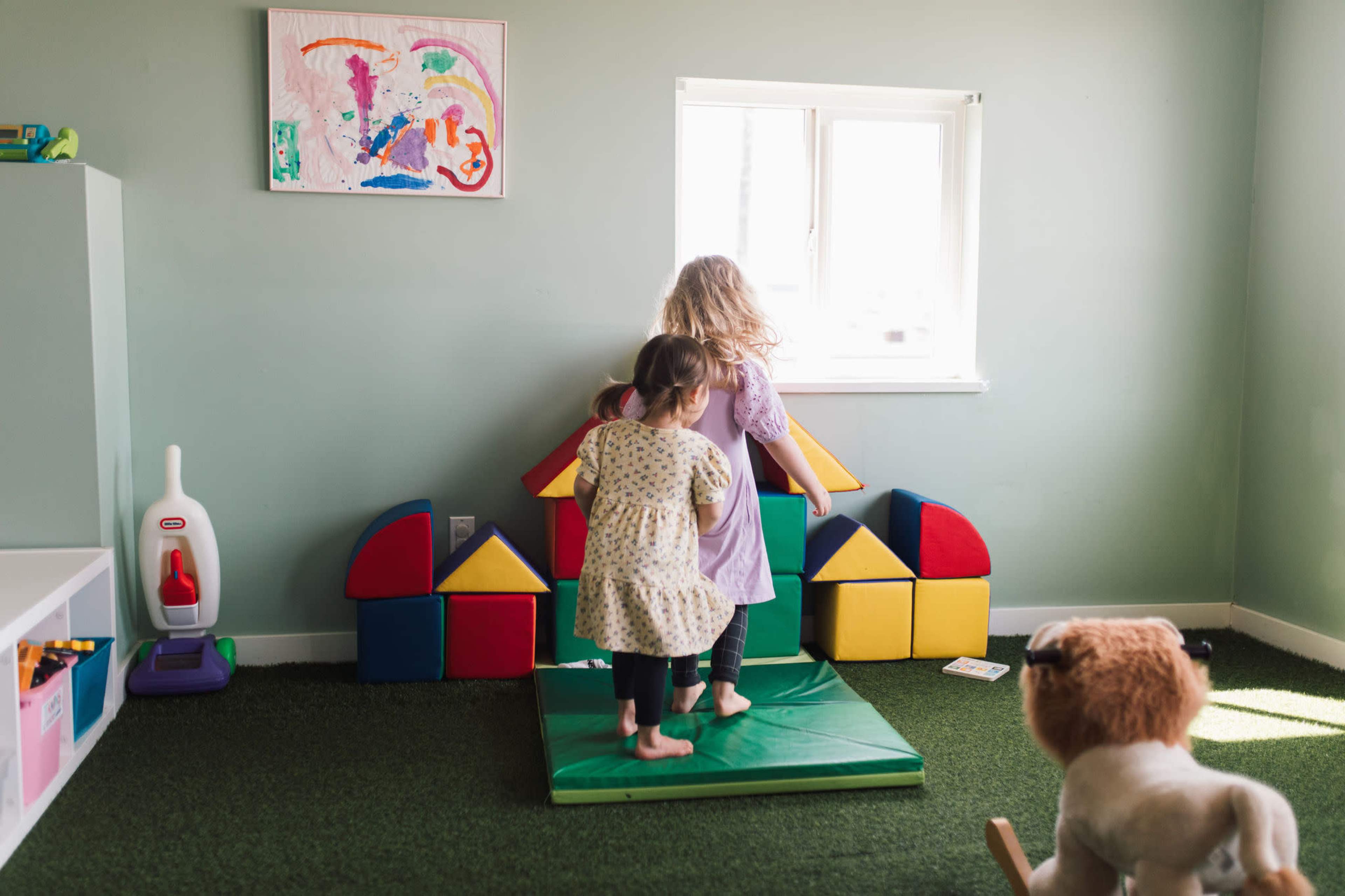 Two children stand on a green mat in a brightly colored playroom with building blocks arranged in the shape of a house and a window in the background.