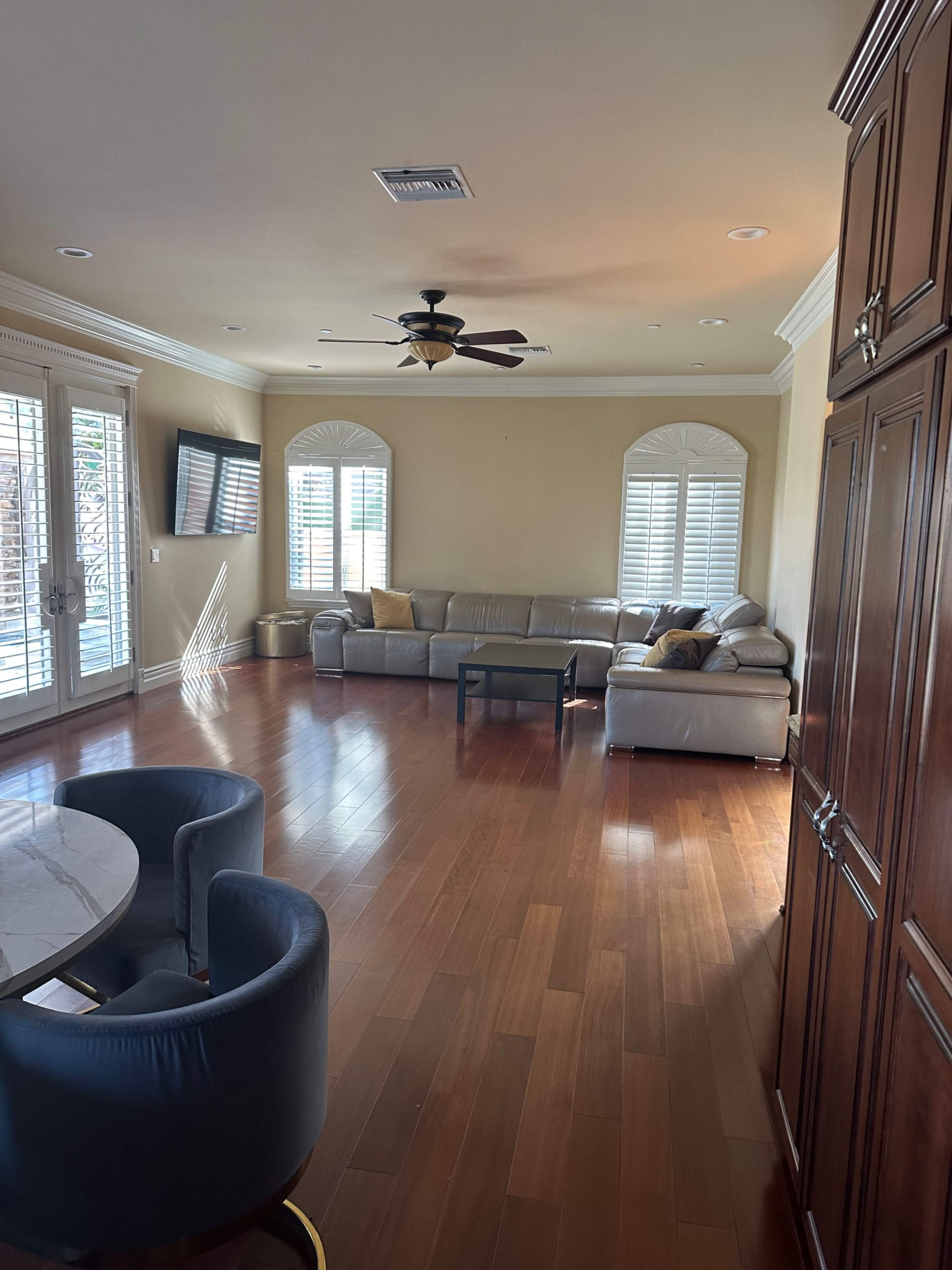 The image shows a spacious living room with a tan leather sectional sofa, wooden flooring, and large windows with shutters.