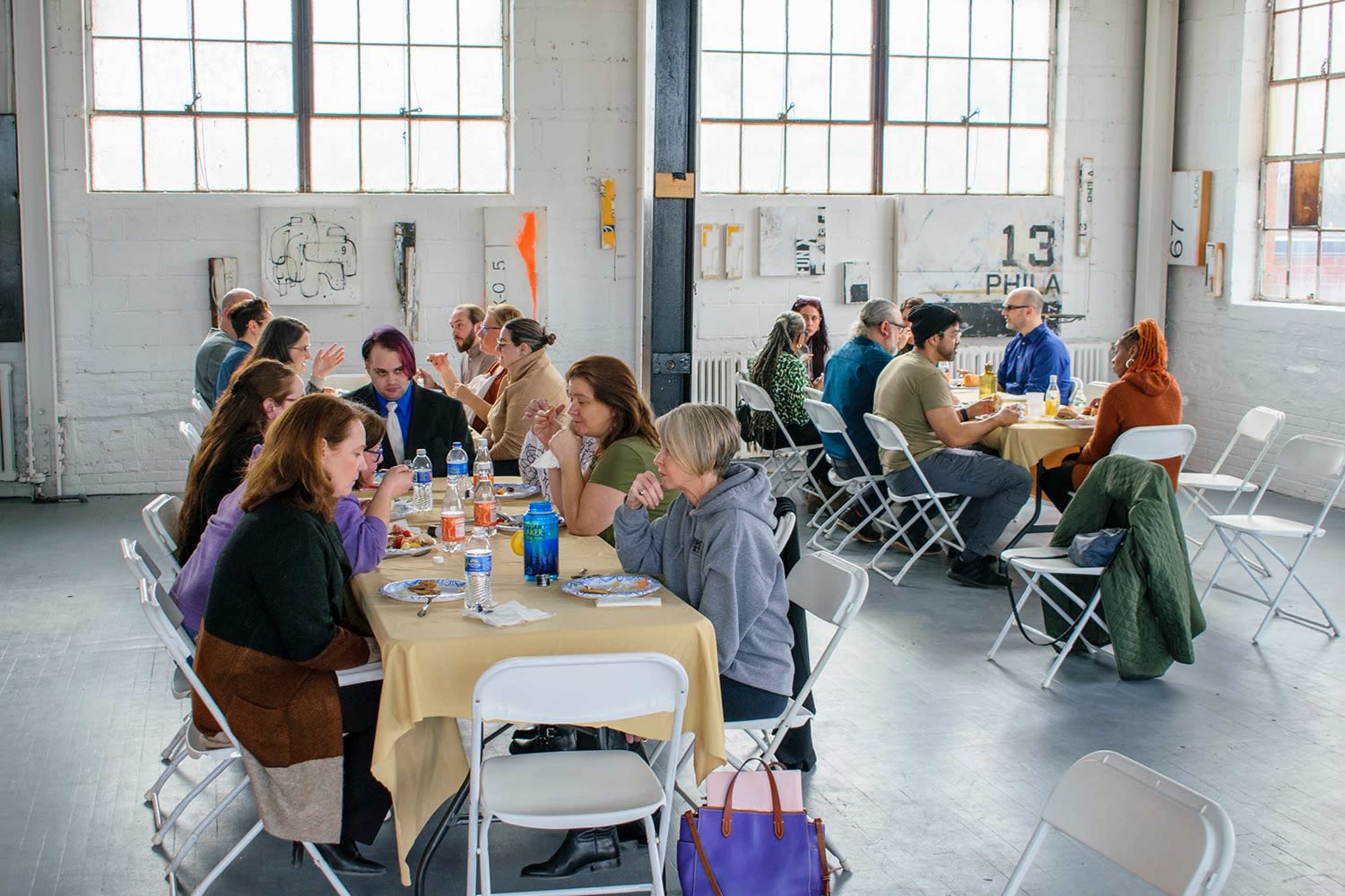 A group of people sits at tables in a spacious room with large windows, eating and conversing during a communal meal.
