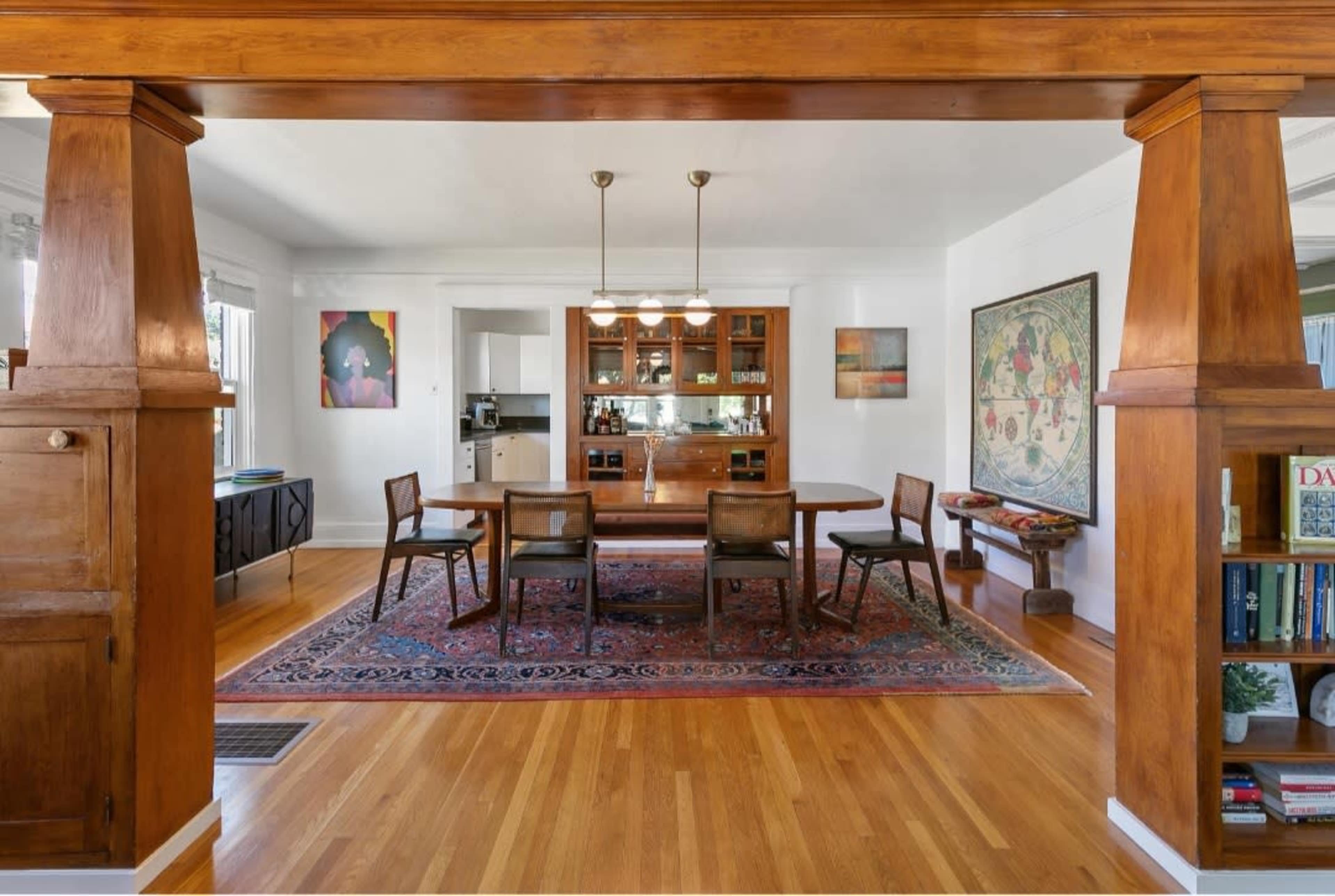 A dining area features a wooden table surrounded by chairs, with a sideboard and a kitchen visible in the background.
