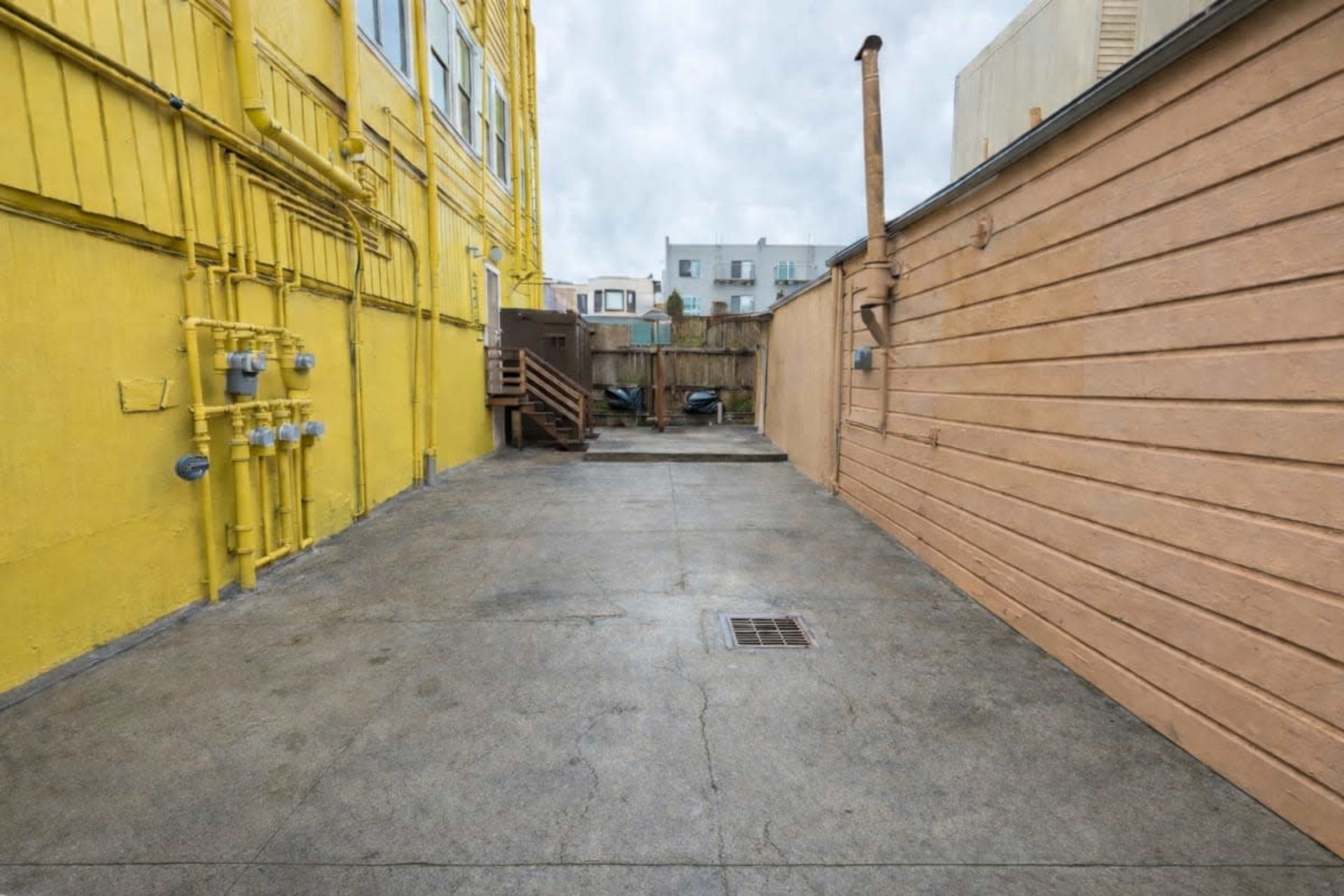 The image shows a narrow, paved alleyway flanked by a bright yellow building on one side and a wooden wall on the other, leading towards a set of stairs and a fenced area in the distance.