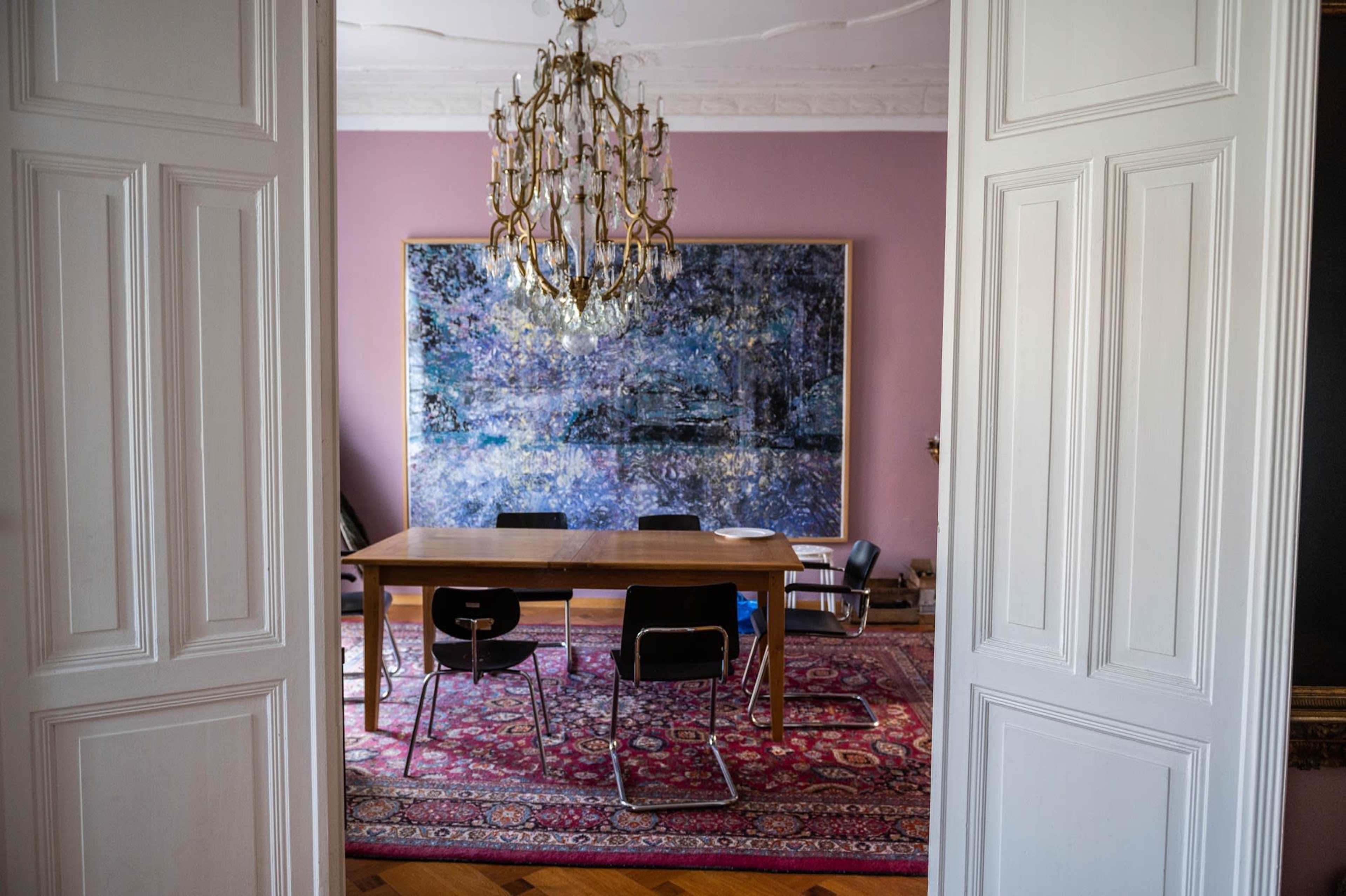 A well-lit dining room with a large painting on the wall and a wooden table surrounded by black chairs, viewed through an open doorway.