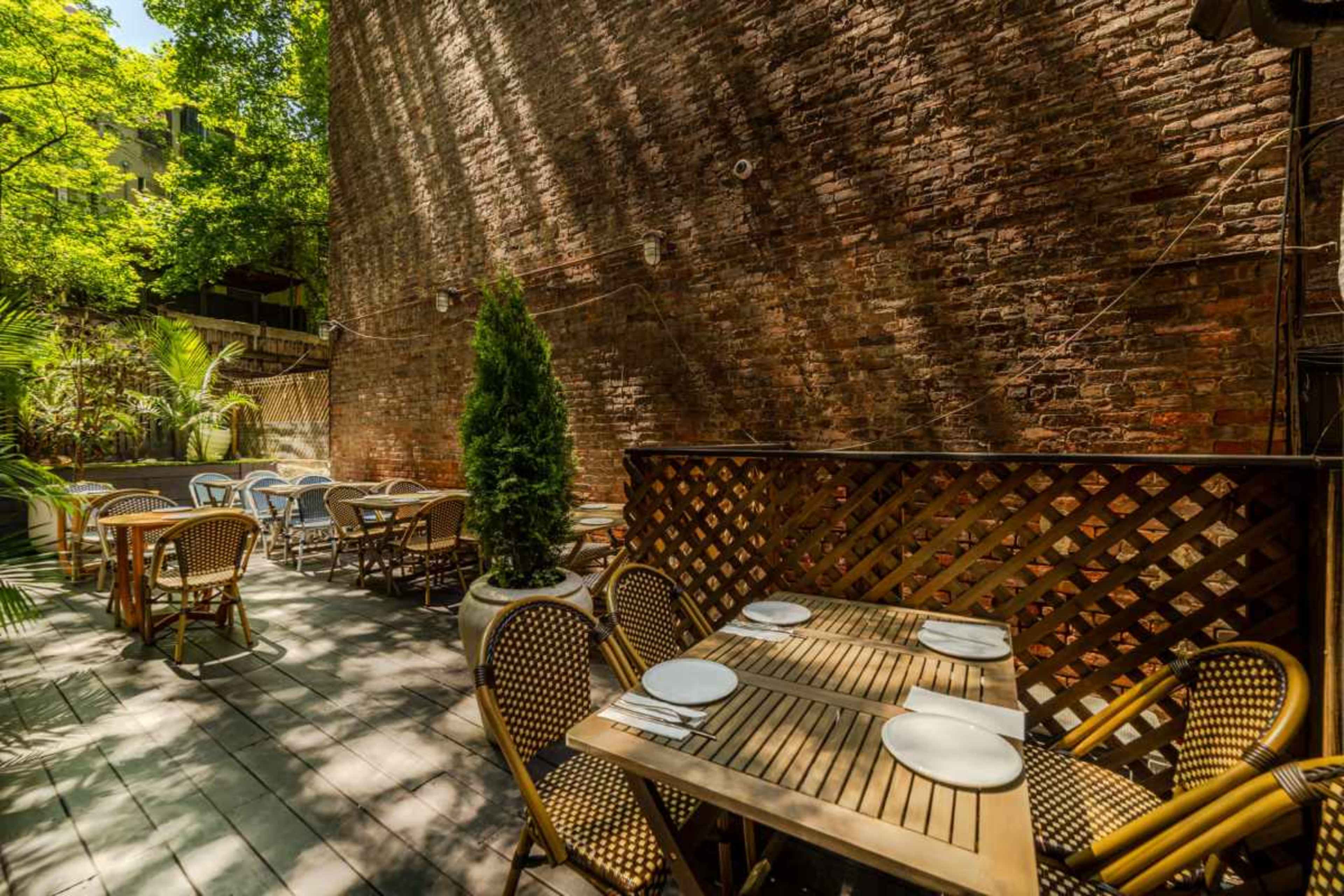 The image shows an outdoor dining area with wooden tables, chairs, and a backdrop of a brick wall and greenery.