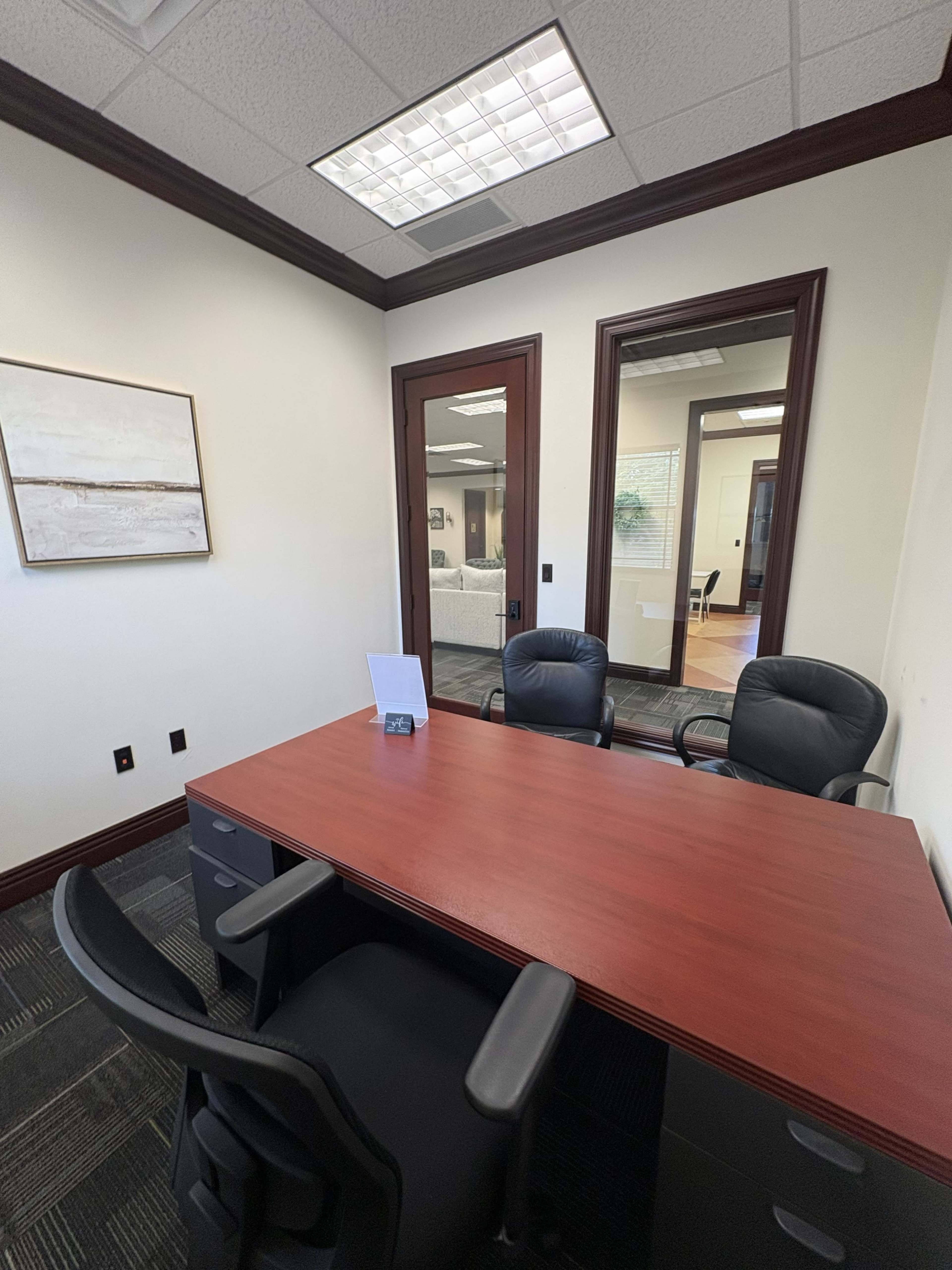 The image shows a small office space featuring a wooden desk with two black chairs, framed artwork on the wall, and a glass door leading to another room.