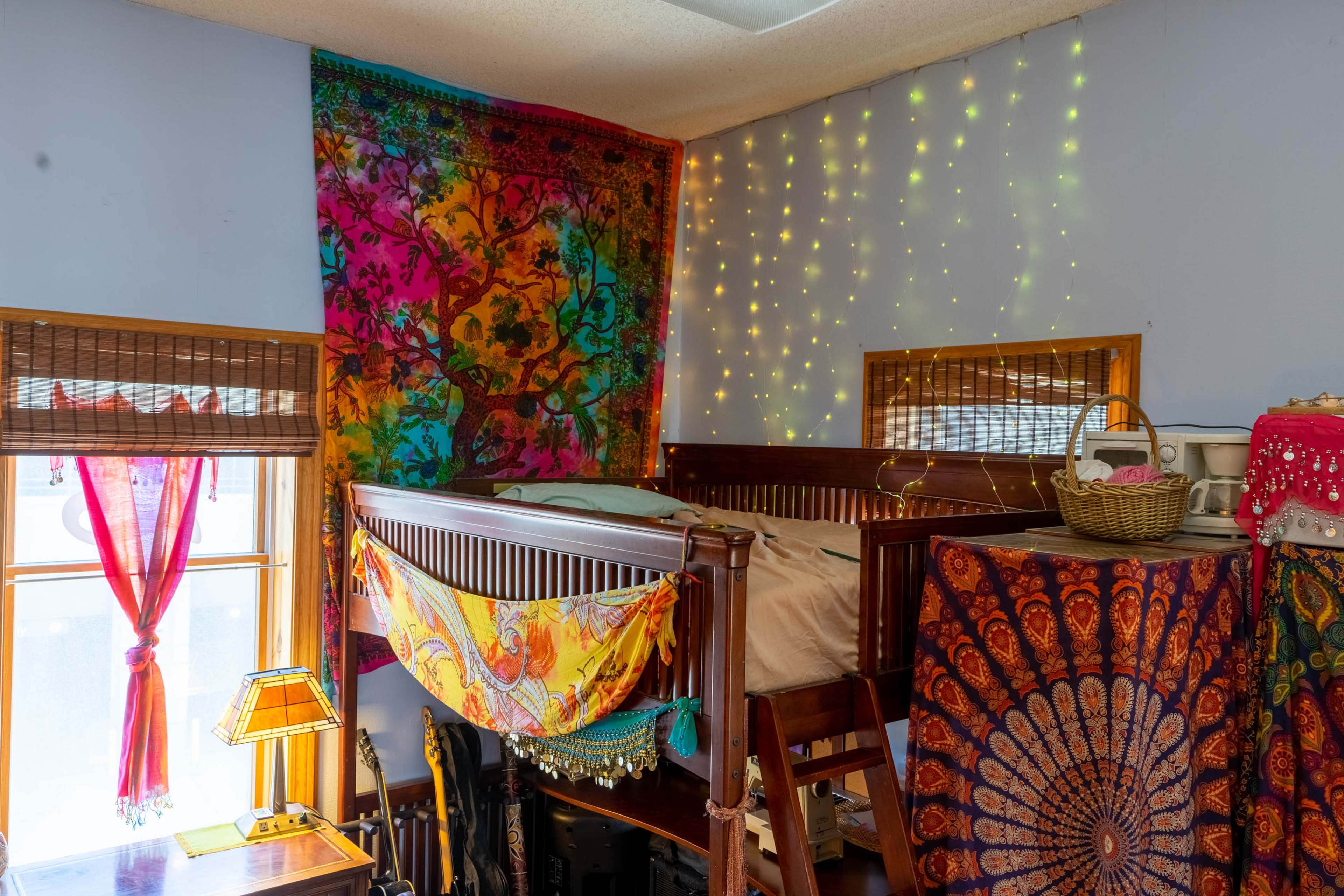 The image shows a colorful bedroom with a lofted bed, decorative tapestries on the walls, and fairy lights creating a cozy atmosphere.