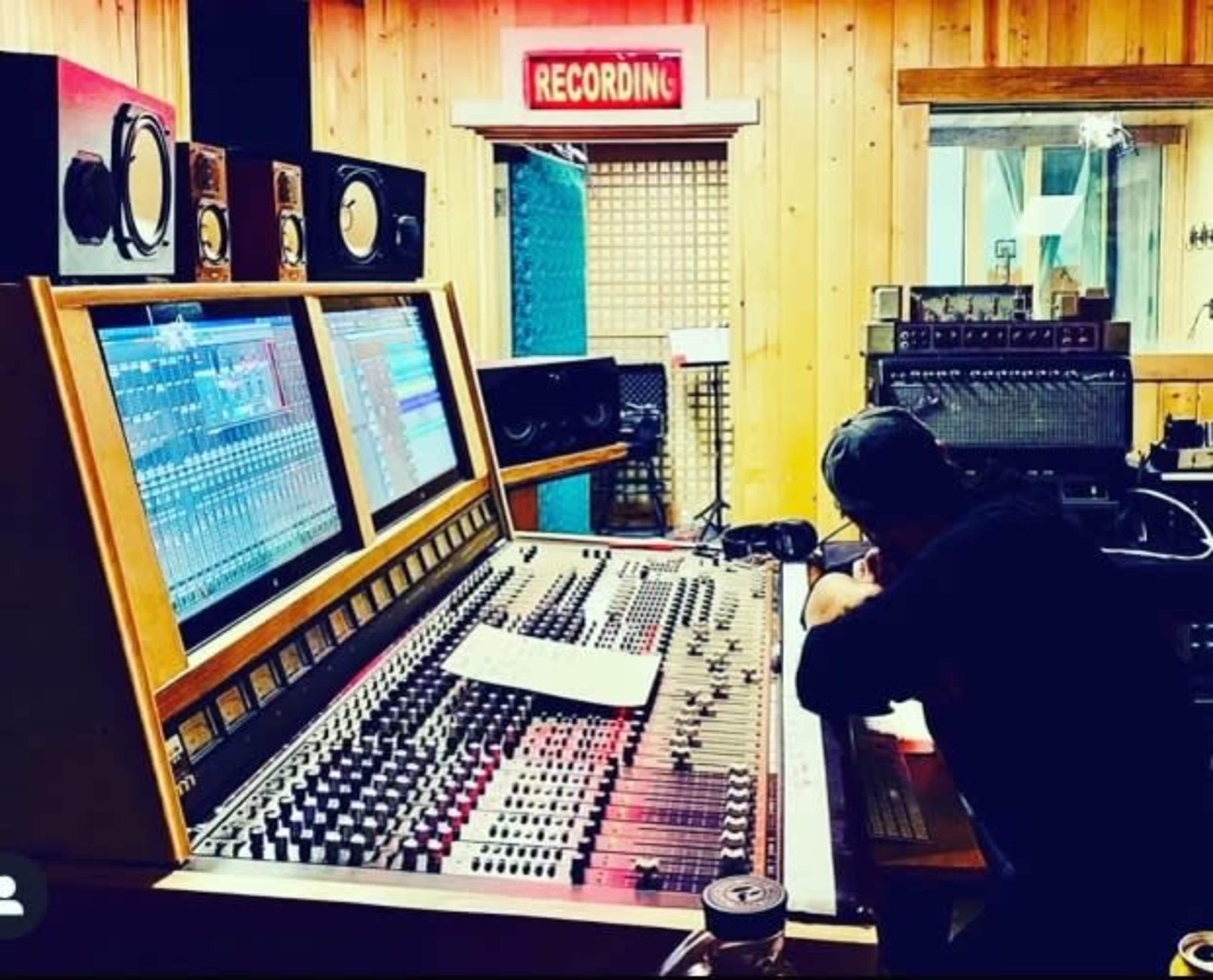 A recording studio features a large mixing console with digital screens, surrounded by various sound equipment, while a person works at the desk.