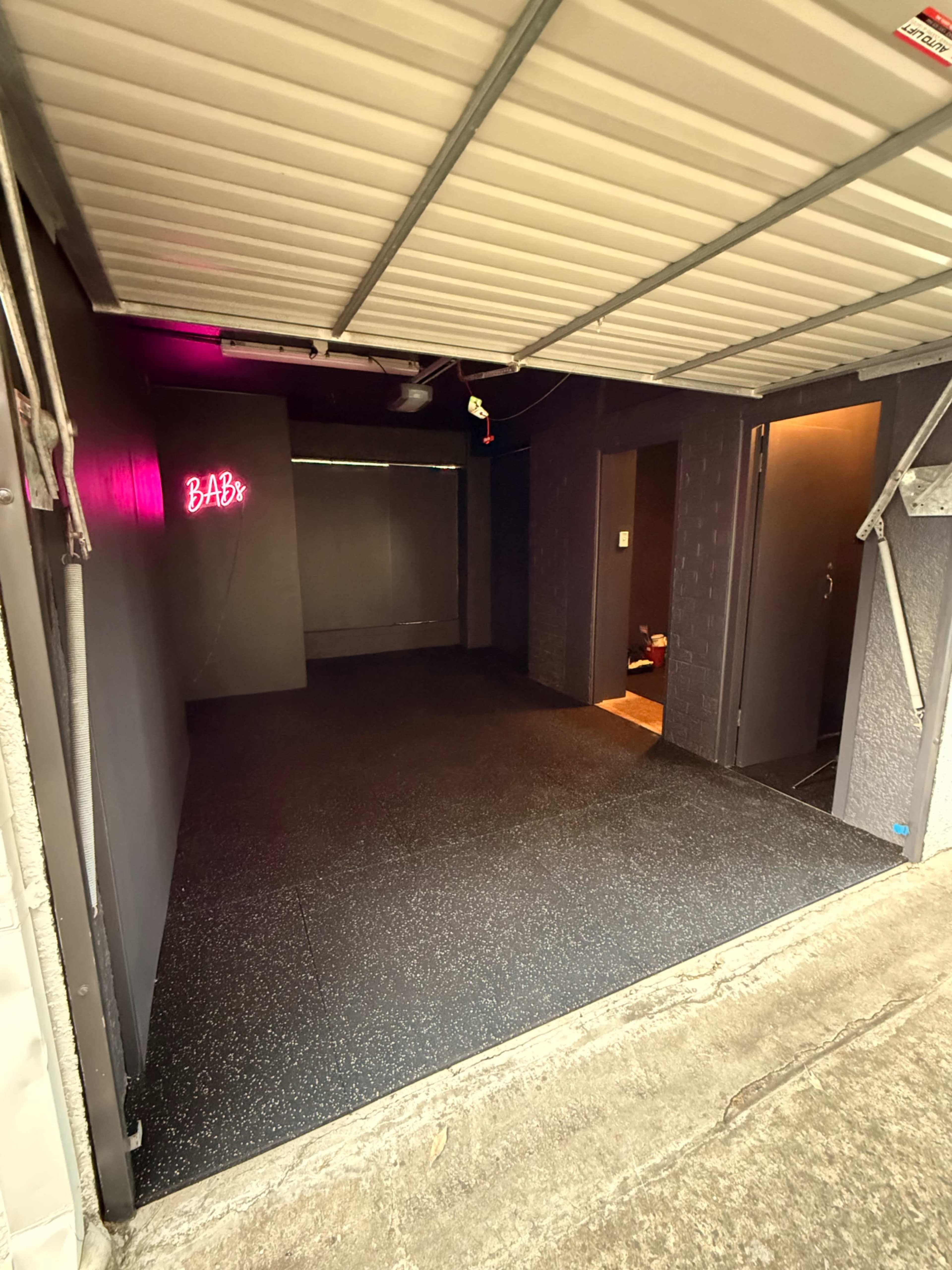 The image shows an entrance to a dimly lit room with dark walls and a black rubber floor, featuring a neon sign that reads "BABY."
