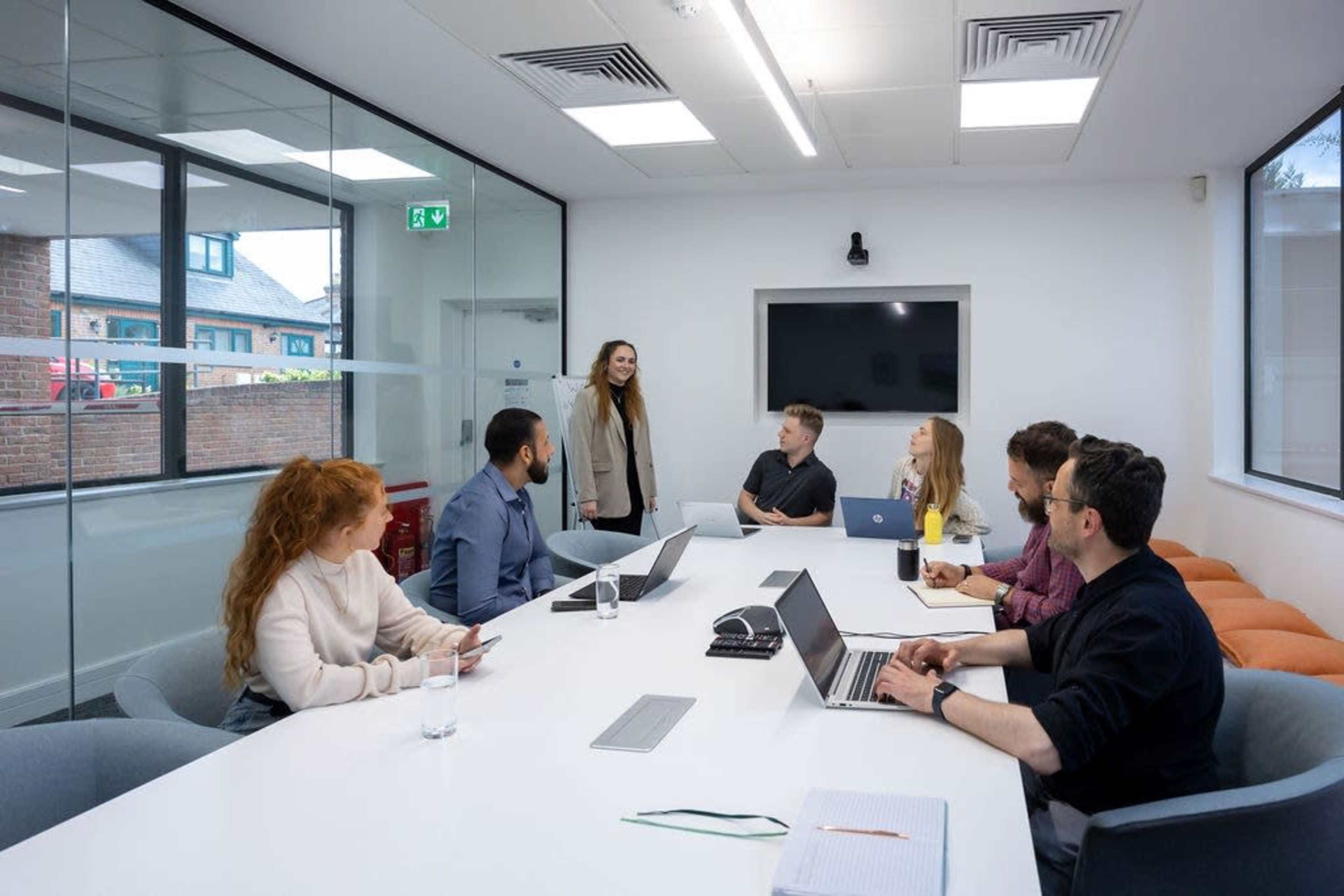 A group of six people are sitting around a long, white conference table in a modern meeting room, while one person stands and addresses the group.