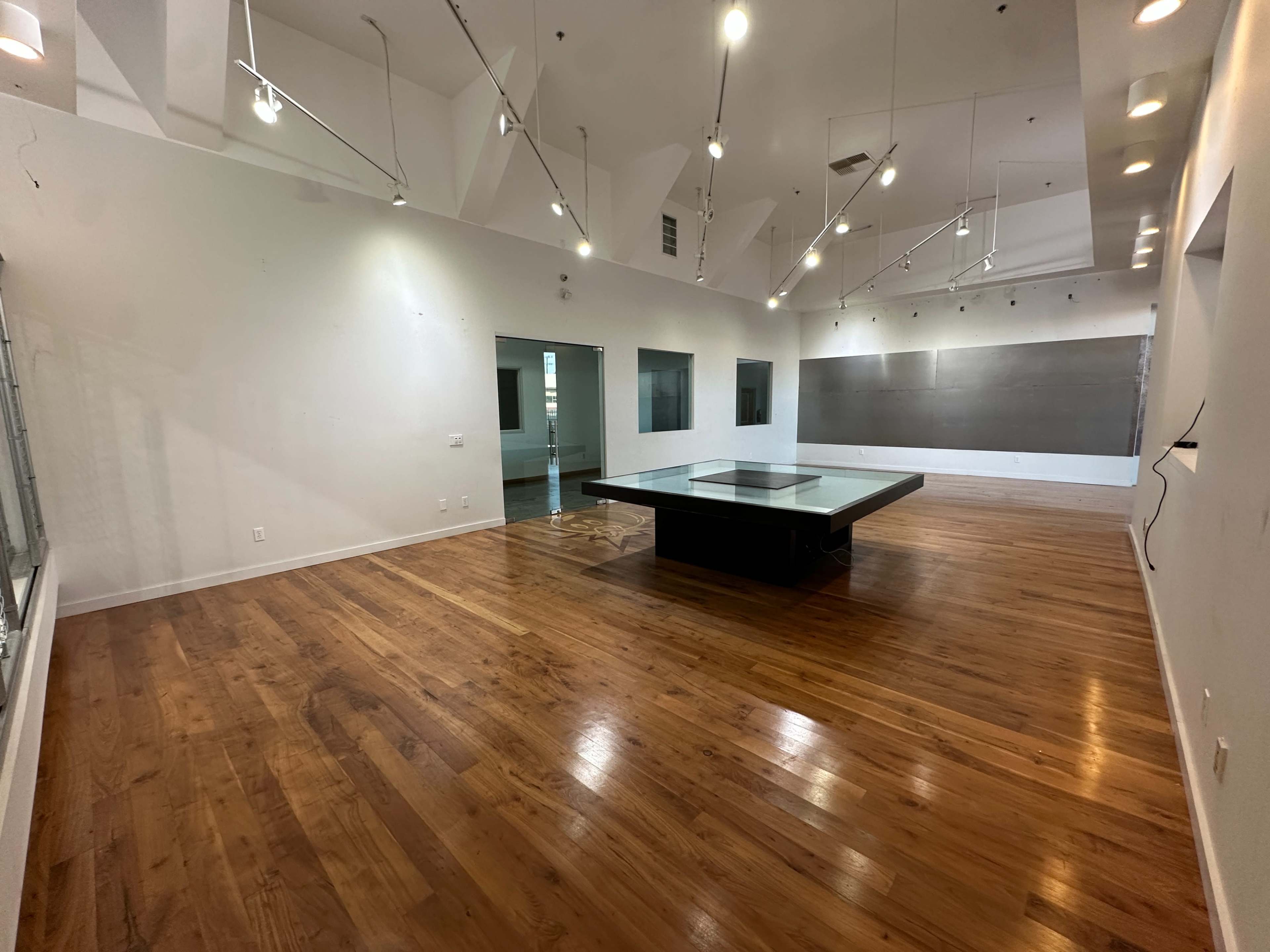 The image shows a spacious, empty room with hardwood floors, a central glass-topped table, and large wall-mounted paintings.