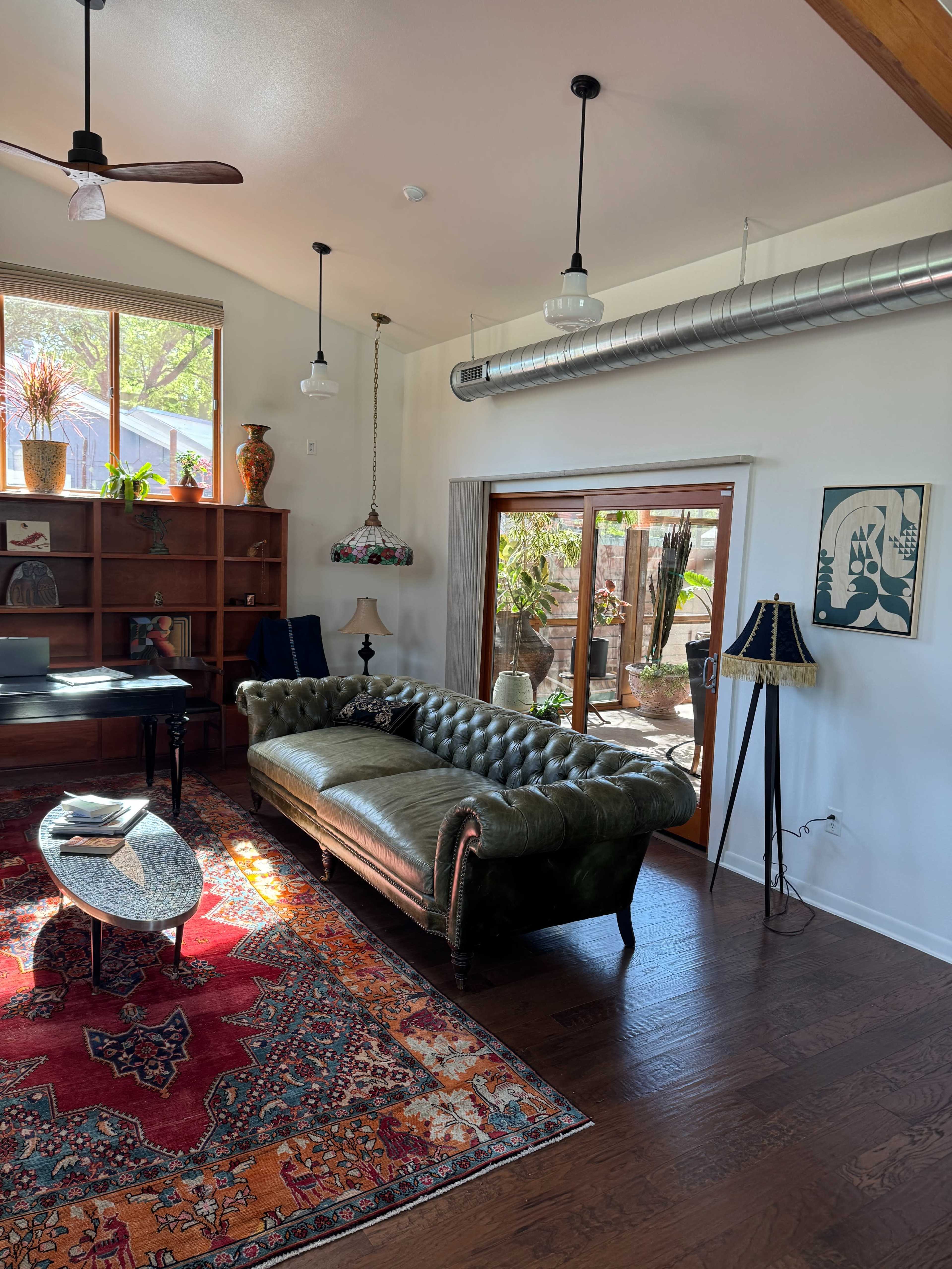 The image shows a living room with a tufted green leather couch, a patterned area rug, a wooden shelf filled with plants, and large windows allowing natural light to illuminate the space.