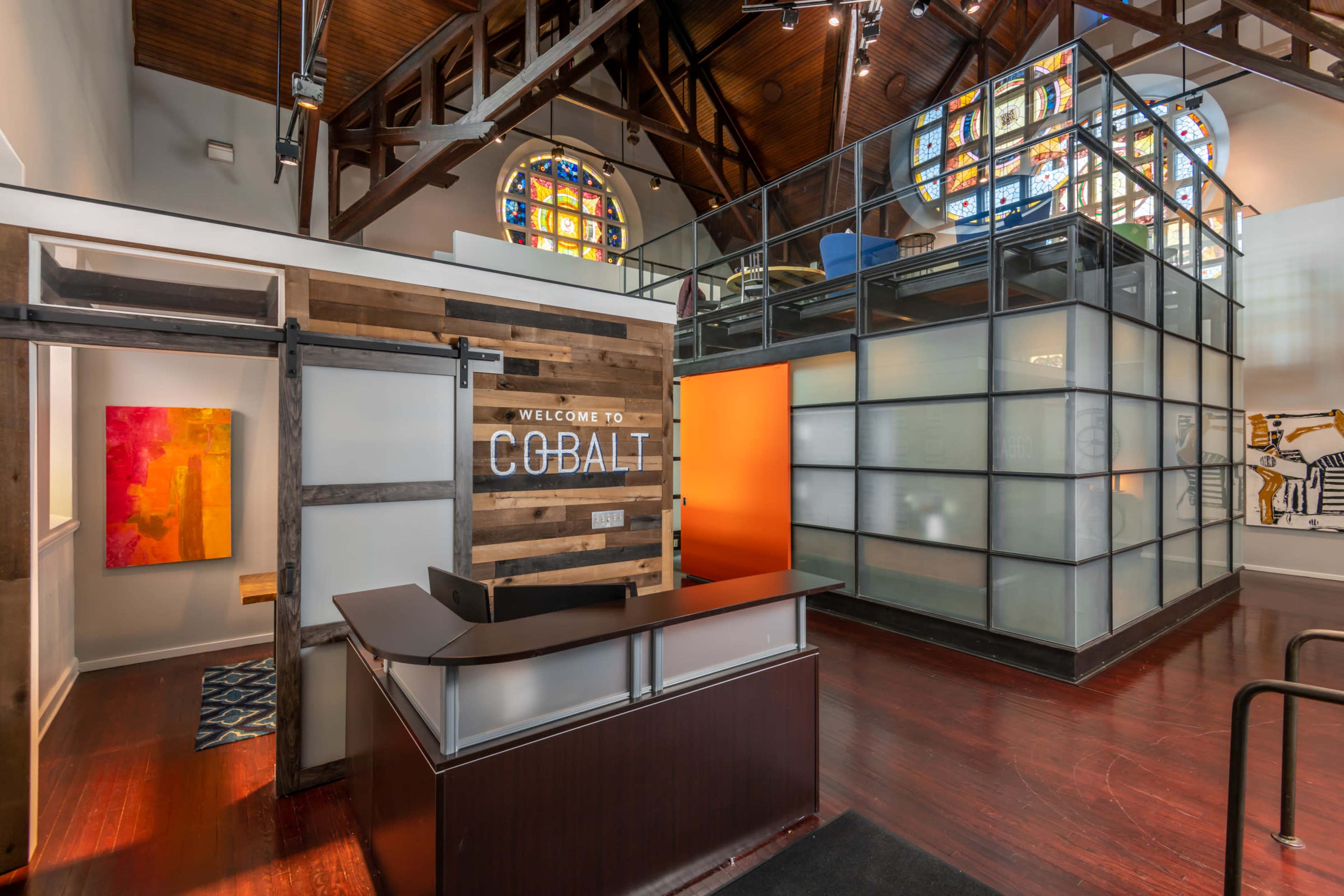 A modern reception area with a wood-paneled desk labeled "WELCOME TO COBALT" and colorful artwork in a spacious room featuring stained glass windows.