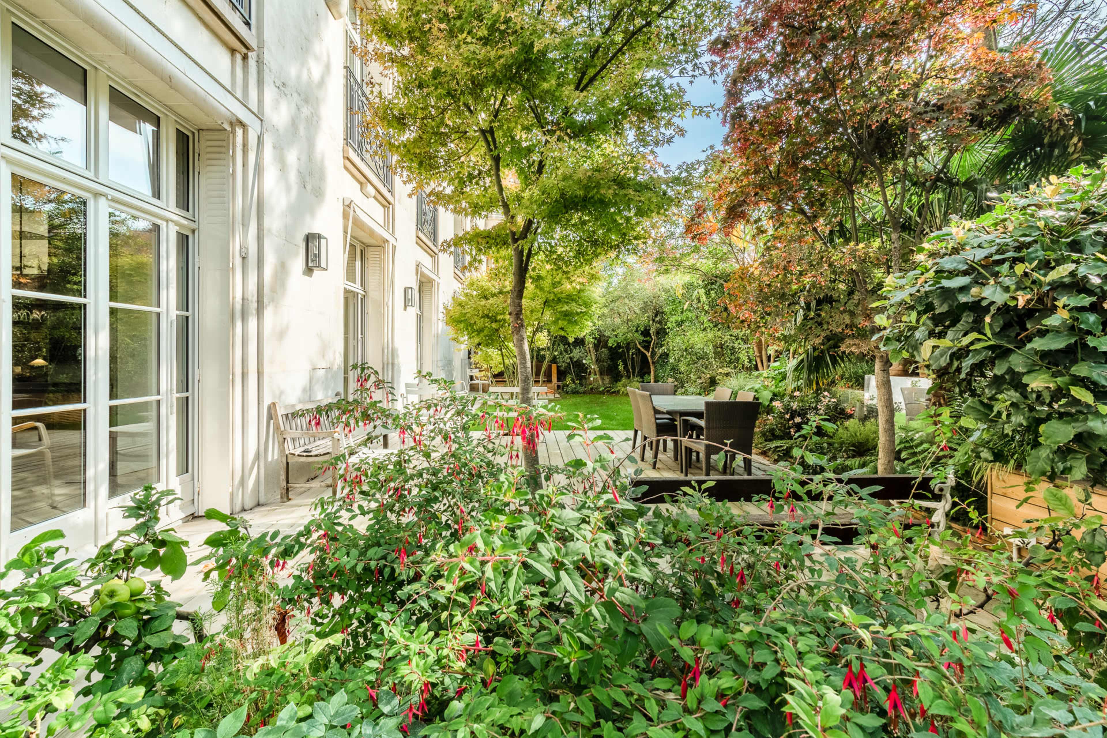A well-maintained garden with green plants and trees beside a building, featuring outdoor seating arrangements.
