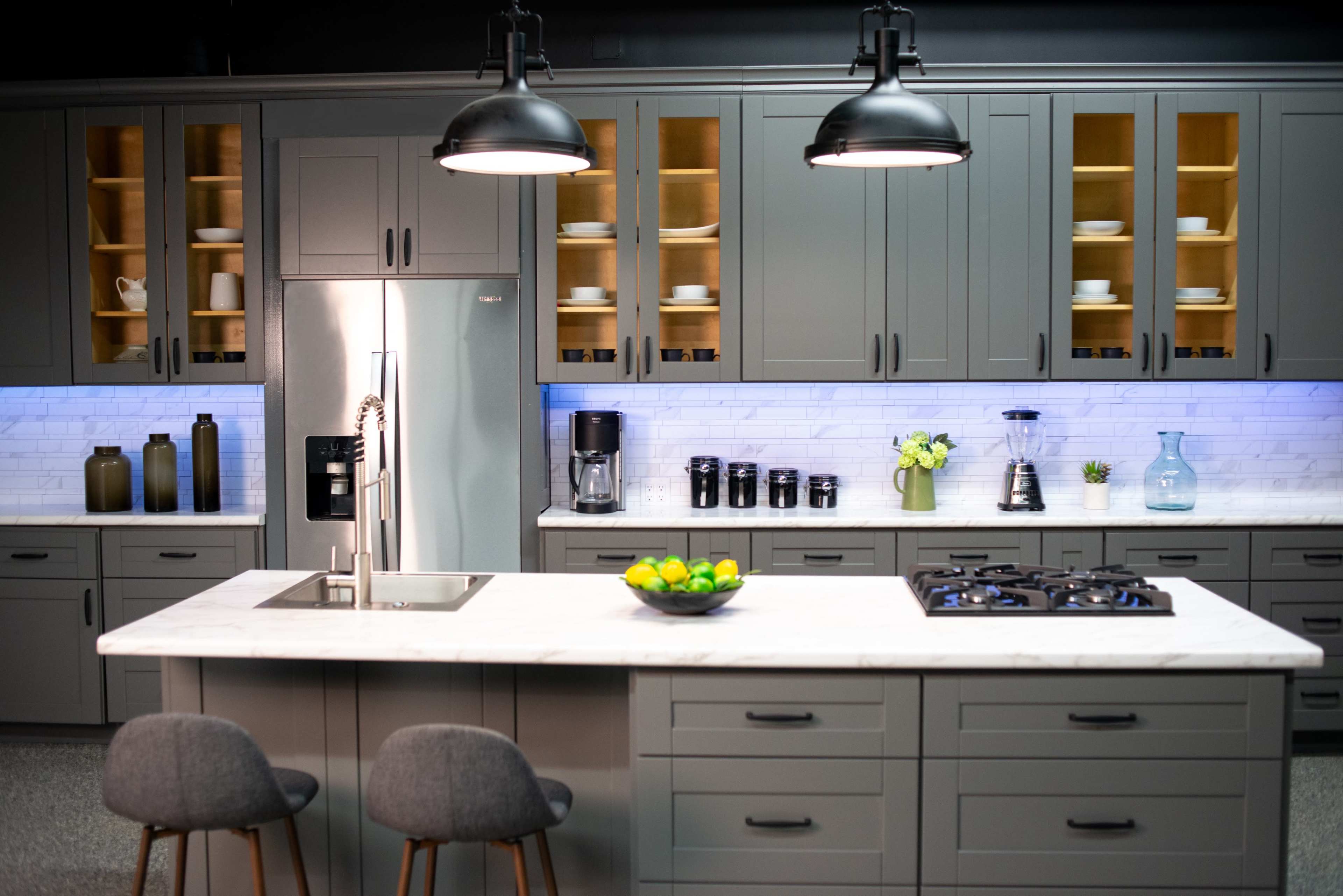 A modern kitchen features gray cabinetry, a marble island with bar stools, and various kitchen appliances against a backdrop of illuminated shelves.
