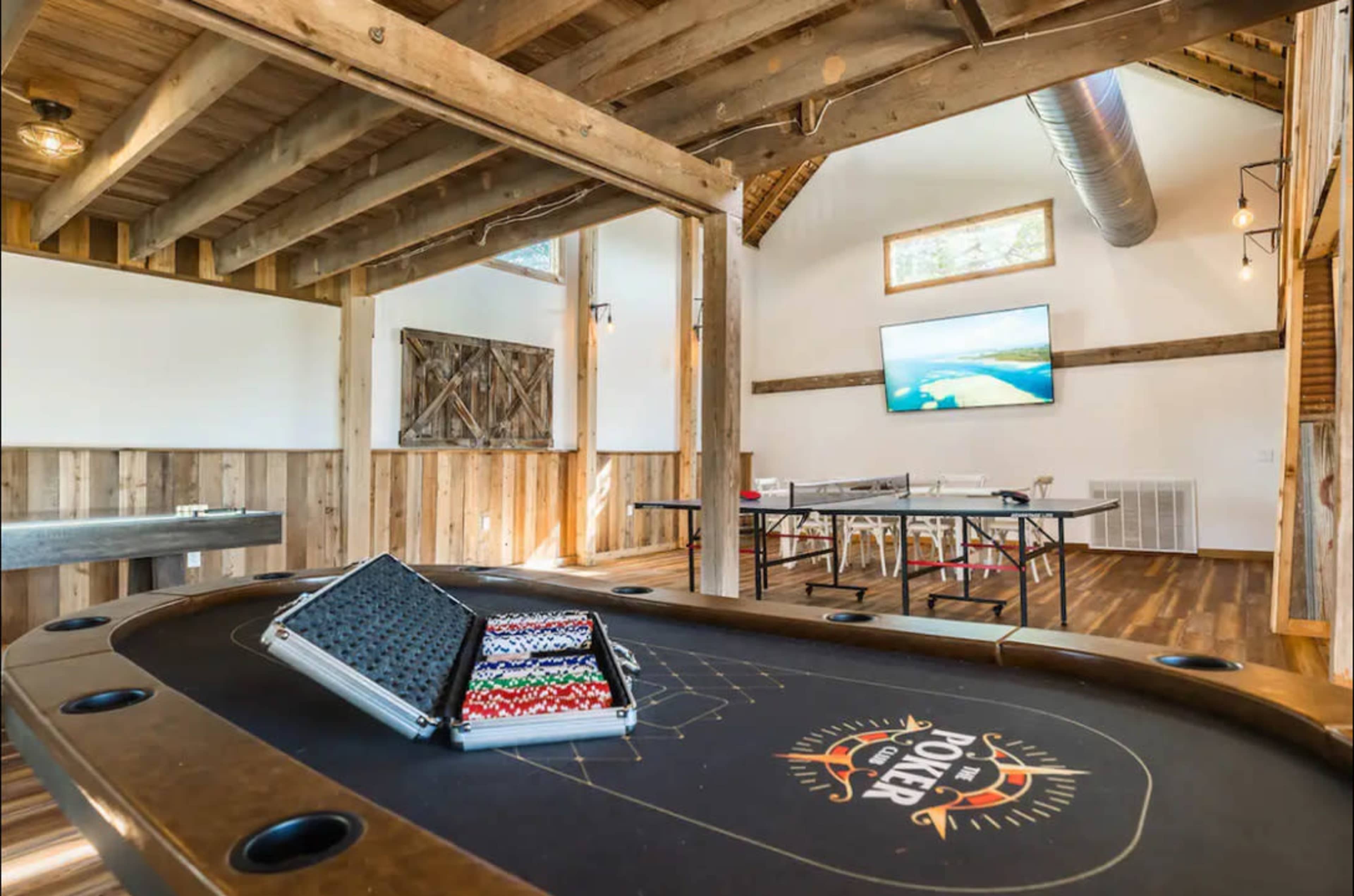 A cozy game room featuring a poker table with chips, wooden beams, and a large wall-mounted screen in a rustic setting.