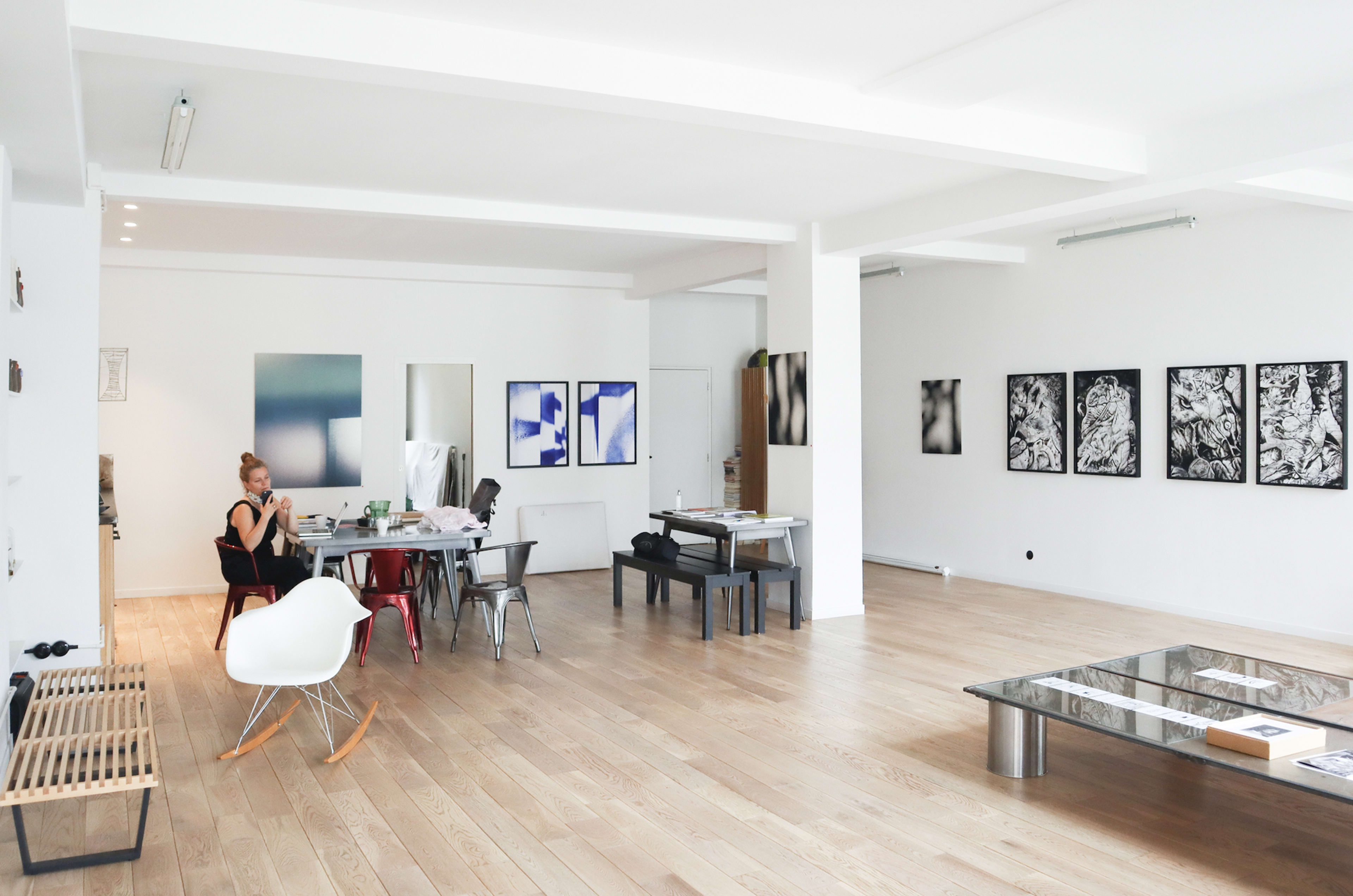 The image shows a spacious, modern art studio with a woman sitting at a table, surrounded by various artworks and furniture.