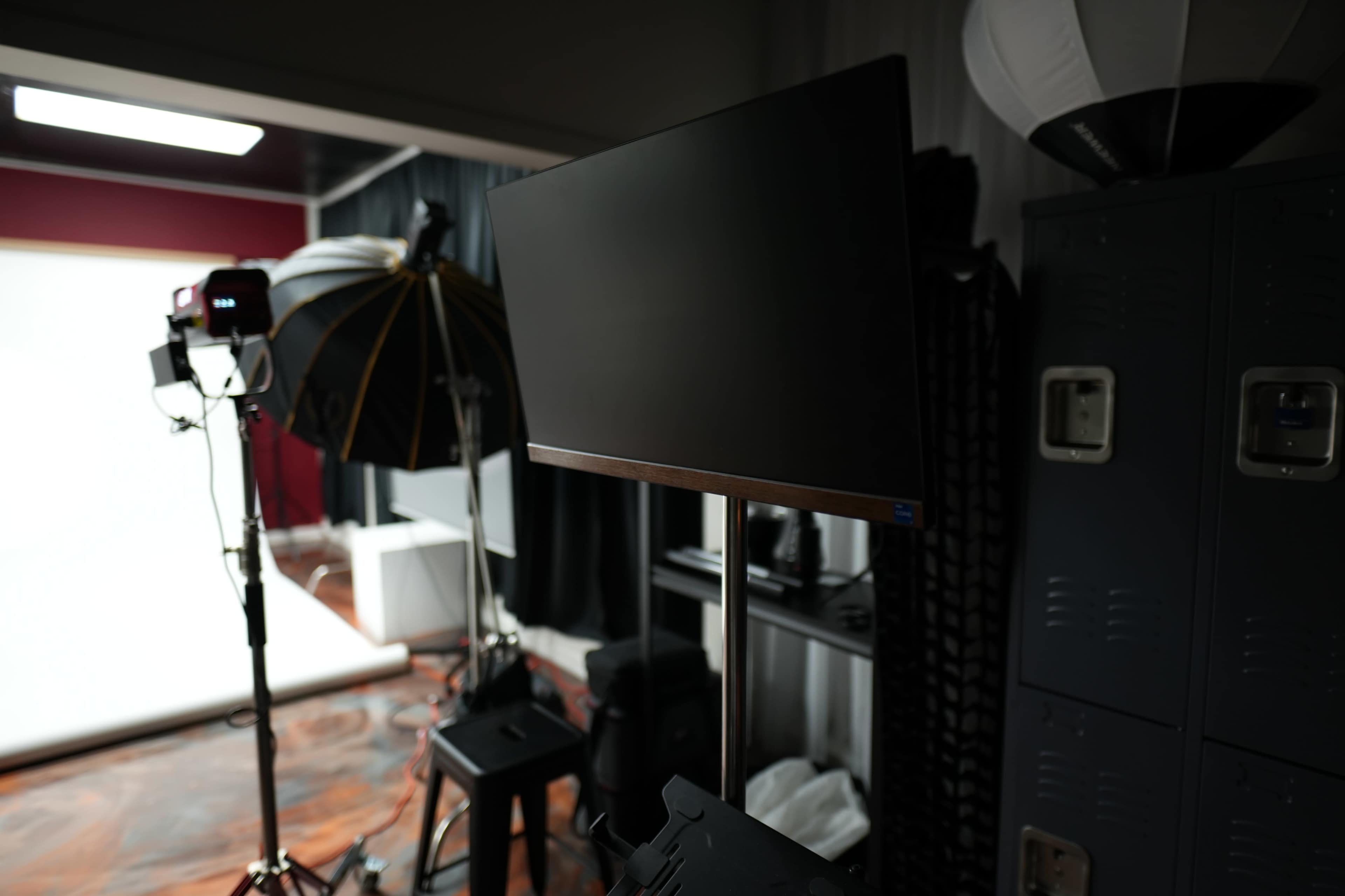 The image shows a photography studio with a monitor on a stand, studio lights, and various equipment along a wall.