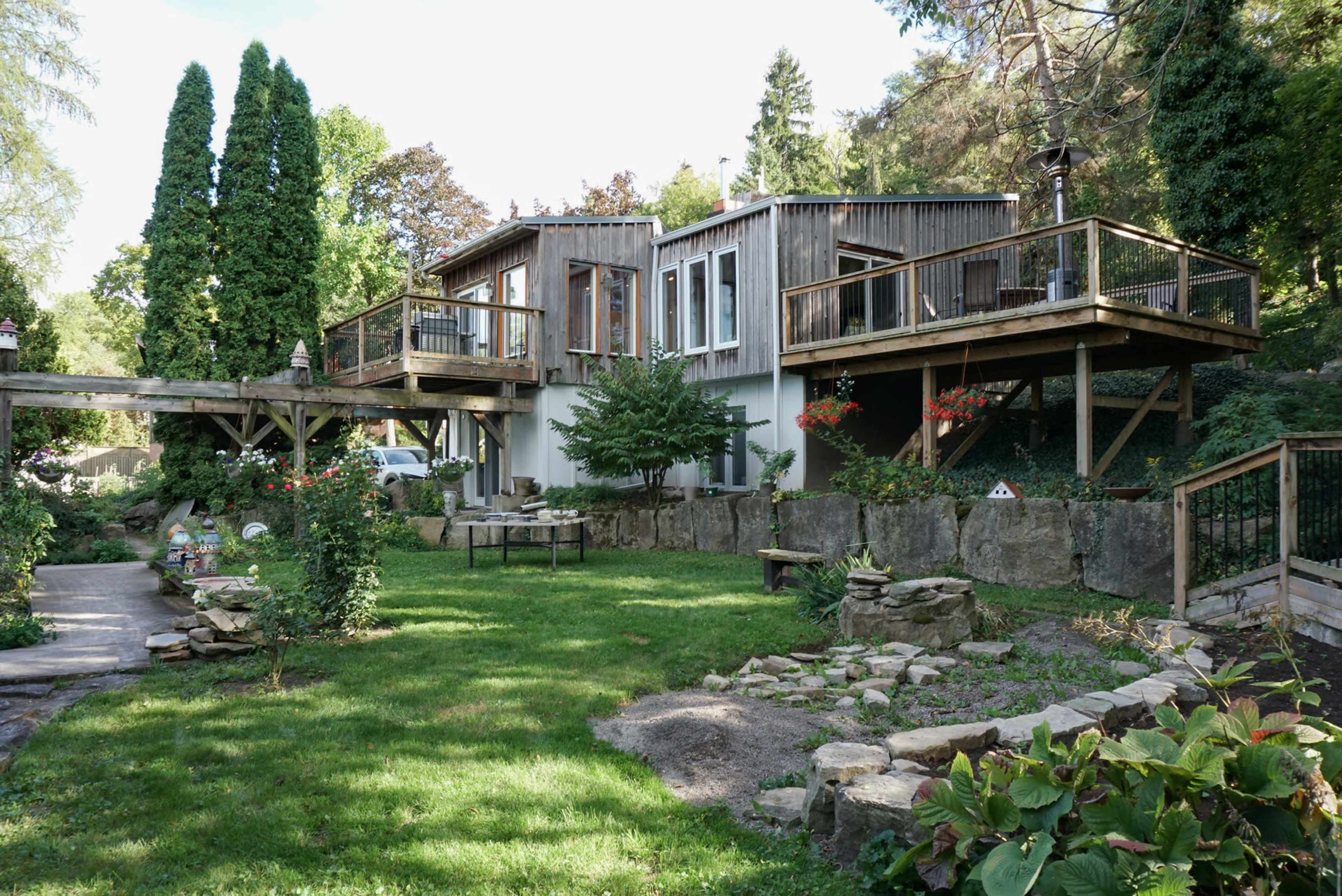 The image depicts a modern house with wooden siding elevated on stilts, surrounded by a lush green lawn and landscaped stone features.