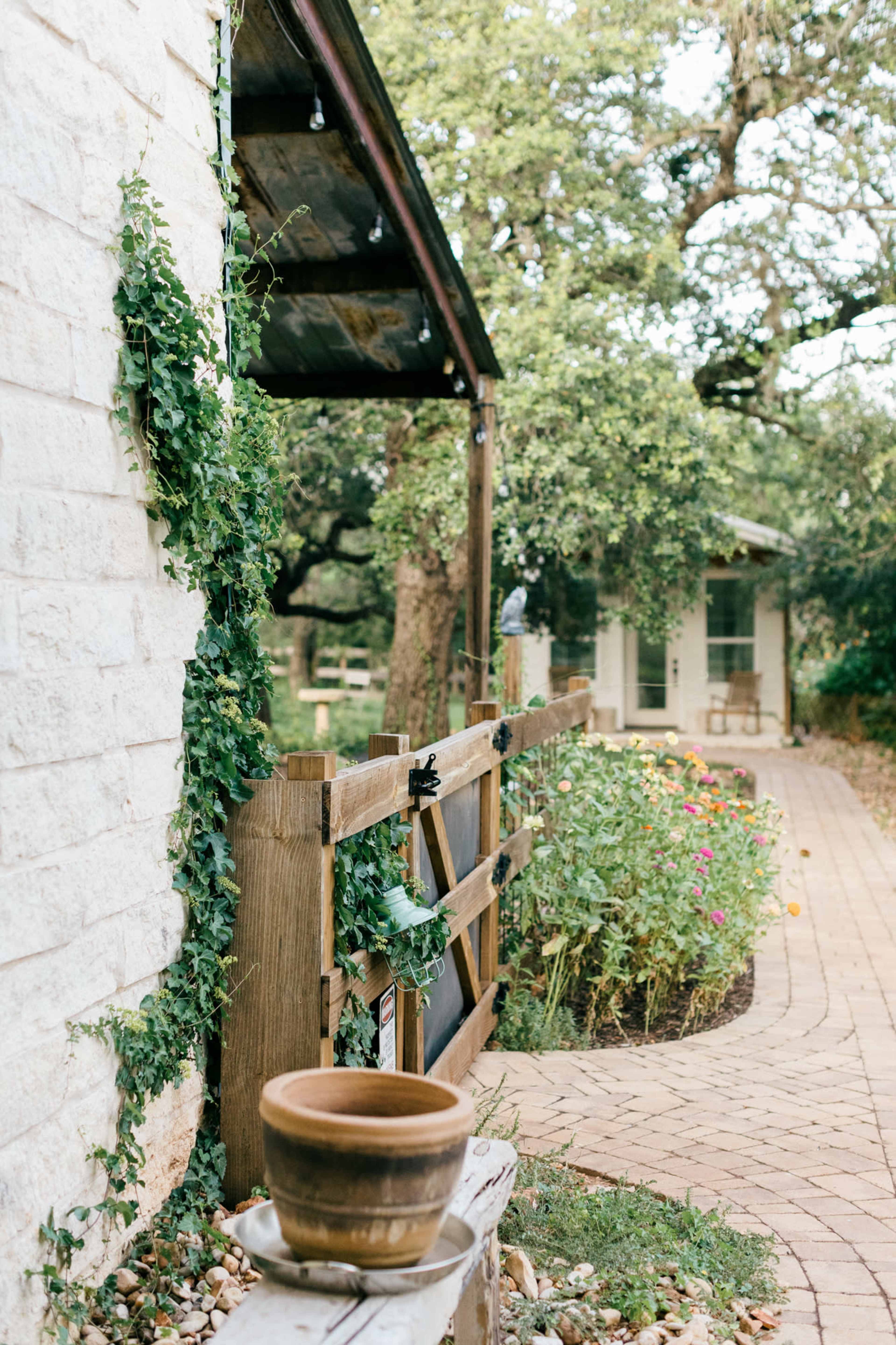 The image shows a rustic wooden gate with a clay pot on a bench, surrounded by lush greenery and a winding path leading to a house.