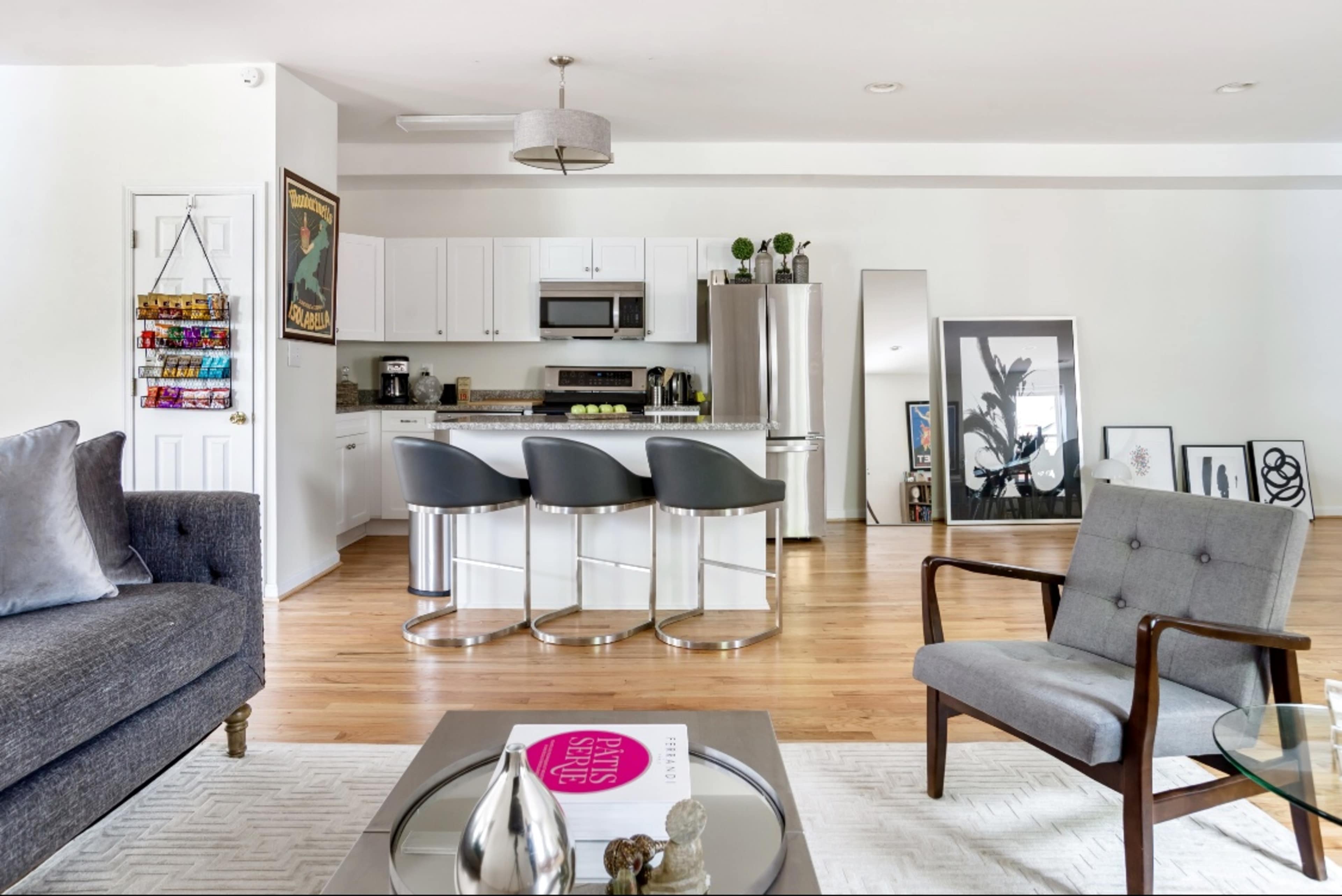 The image shows a modern living room with a gray sofa and armchair, a coffee table with a magazine, and a kitchen area featuring white cabinets and bar stools.