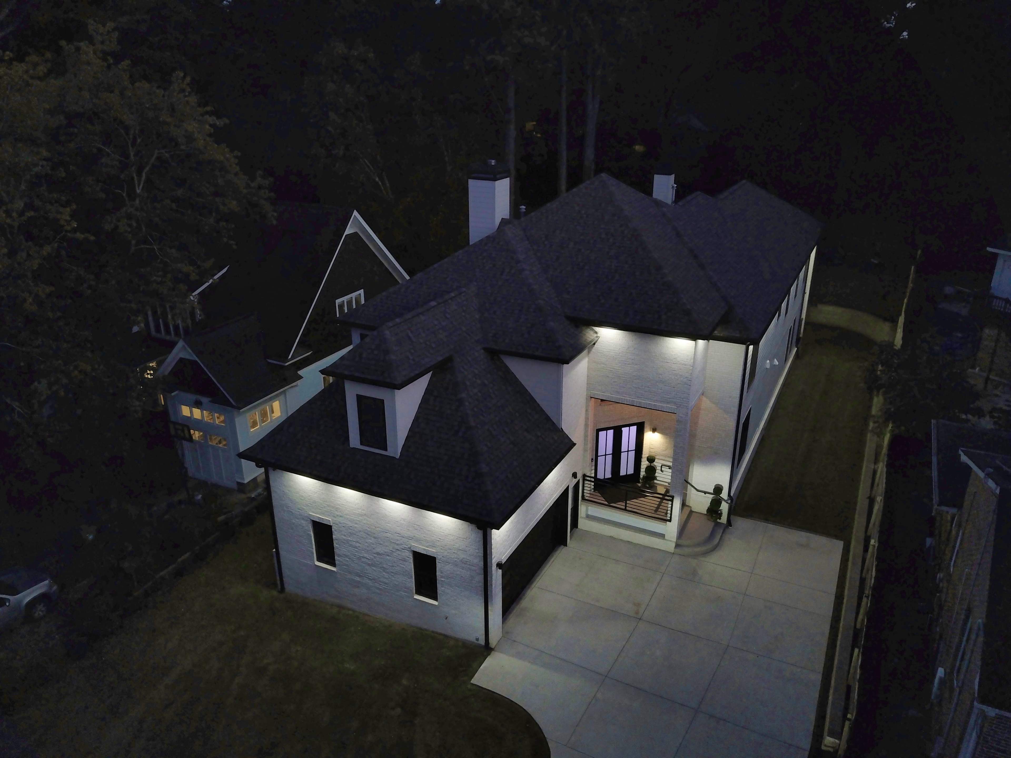 A modern two-story house with a dark roof and white exterior is illuminated by outdoor lights, situated in a wooded area at dusk.