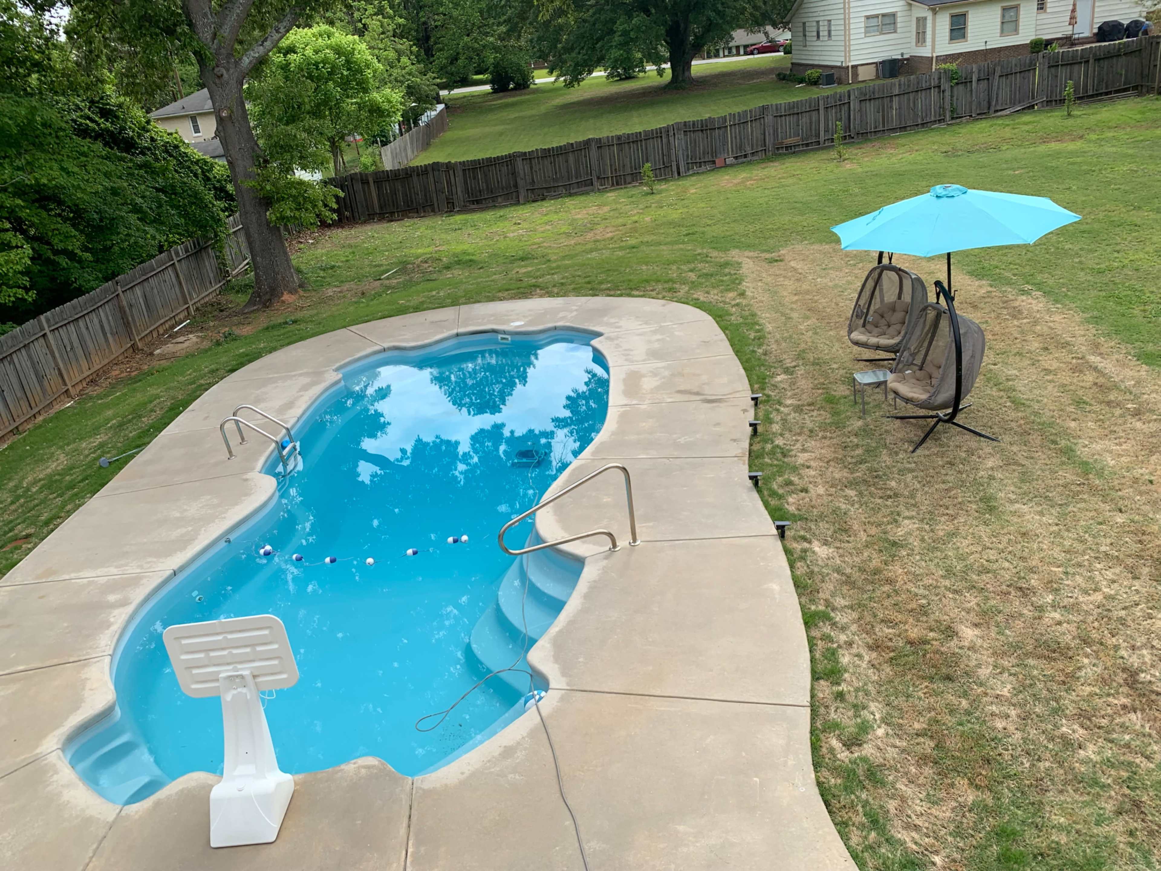 A swimming pool with steps and a diving board is surrounded by a yard featuring two hanging chairs and an umbrella.