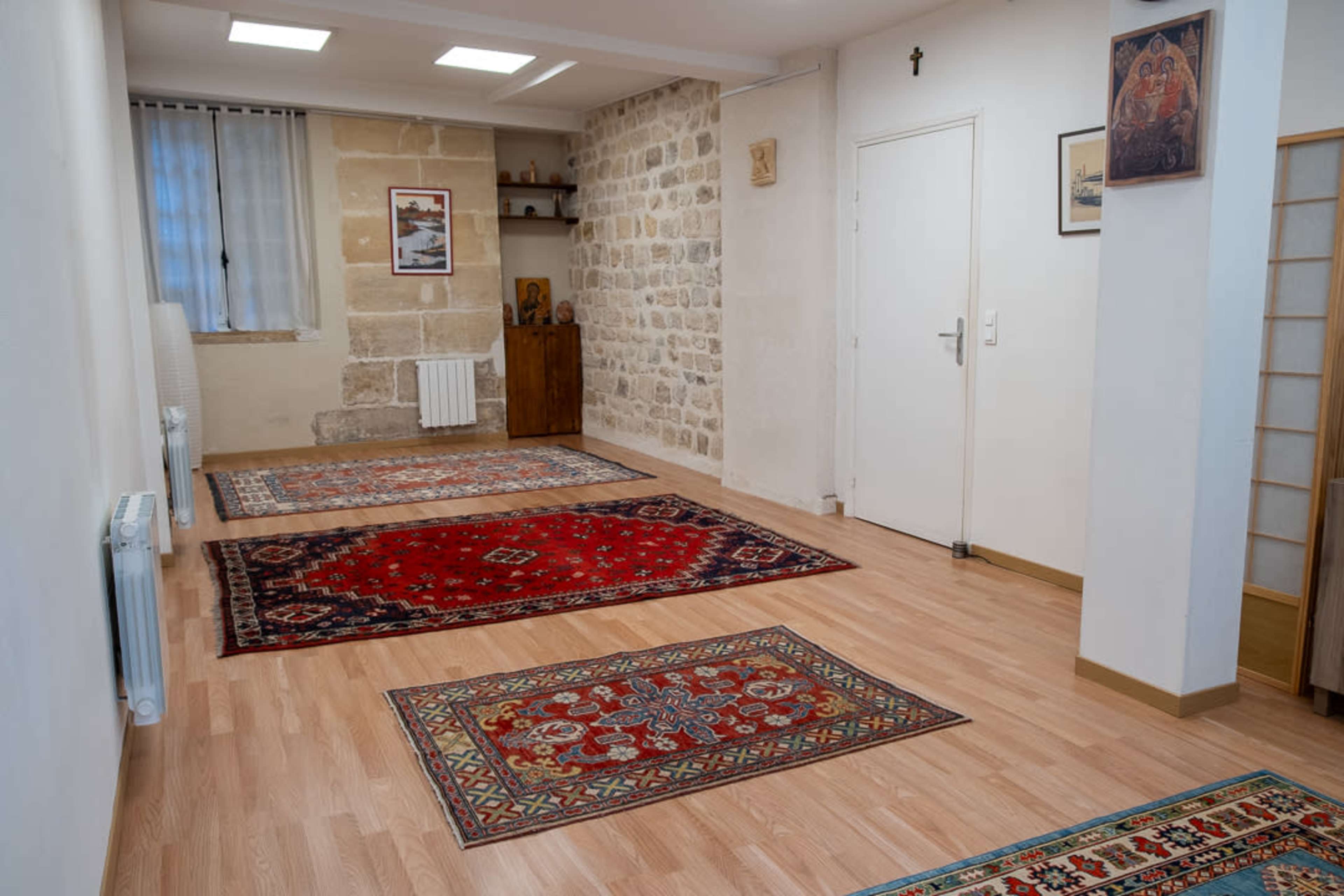 The image shows a spacious, well-lit room featuring wooden flooring and several patterned carpets, with exposed stone walls on one side and a wooden cabinet against the wall.
