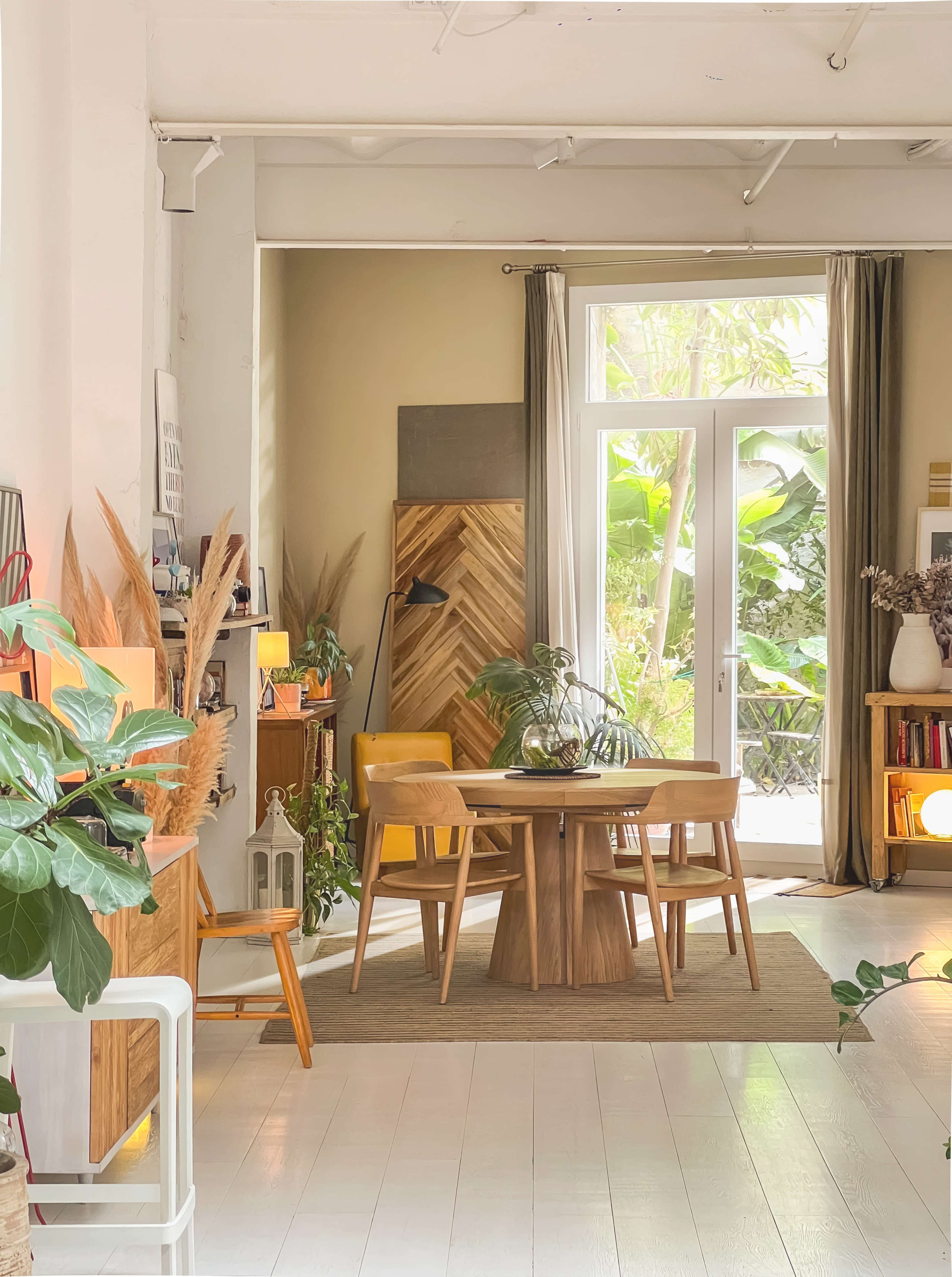 The image shows a brightly lit, modern interior of a dining area with a wooden table and chairs, surrounded by various plants and decor, and featuring large windows that provide a view of greenery outside.