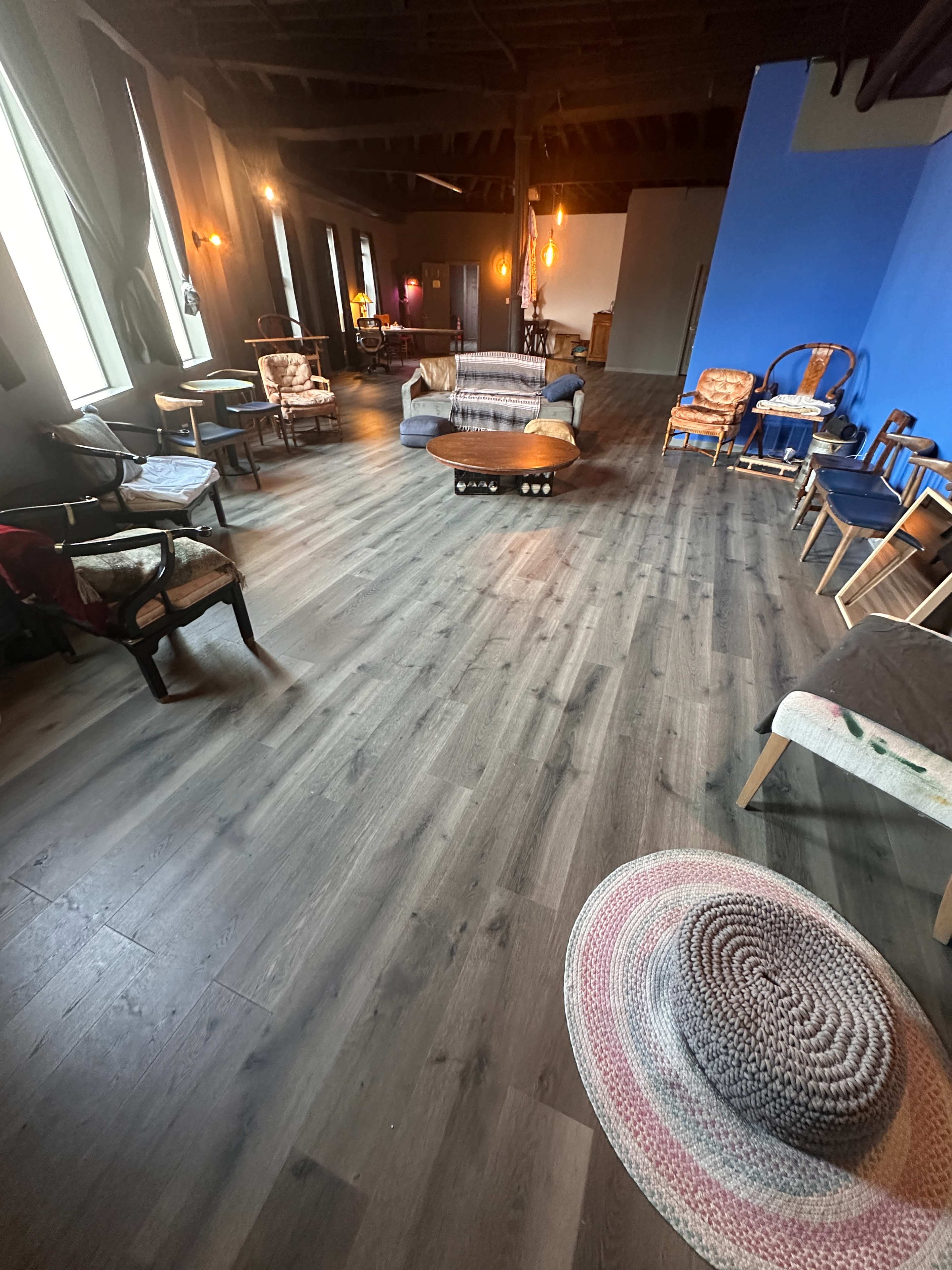The image shows a spacious, well-lit room with wooden flooring, featuring various chairs and couches arranged around a central seating area.