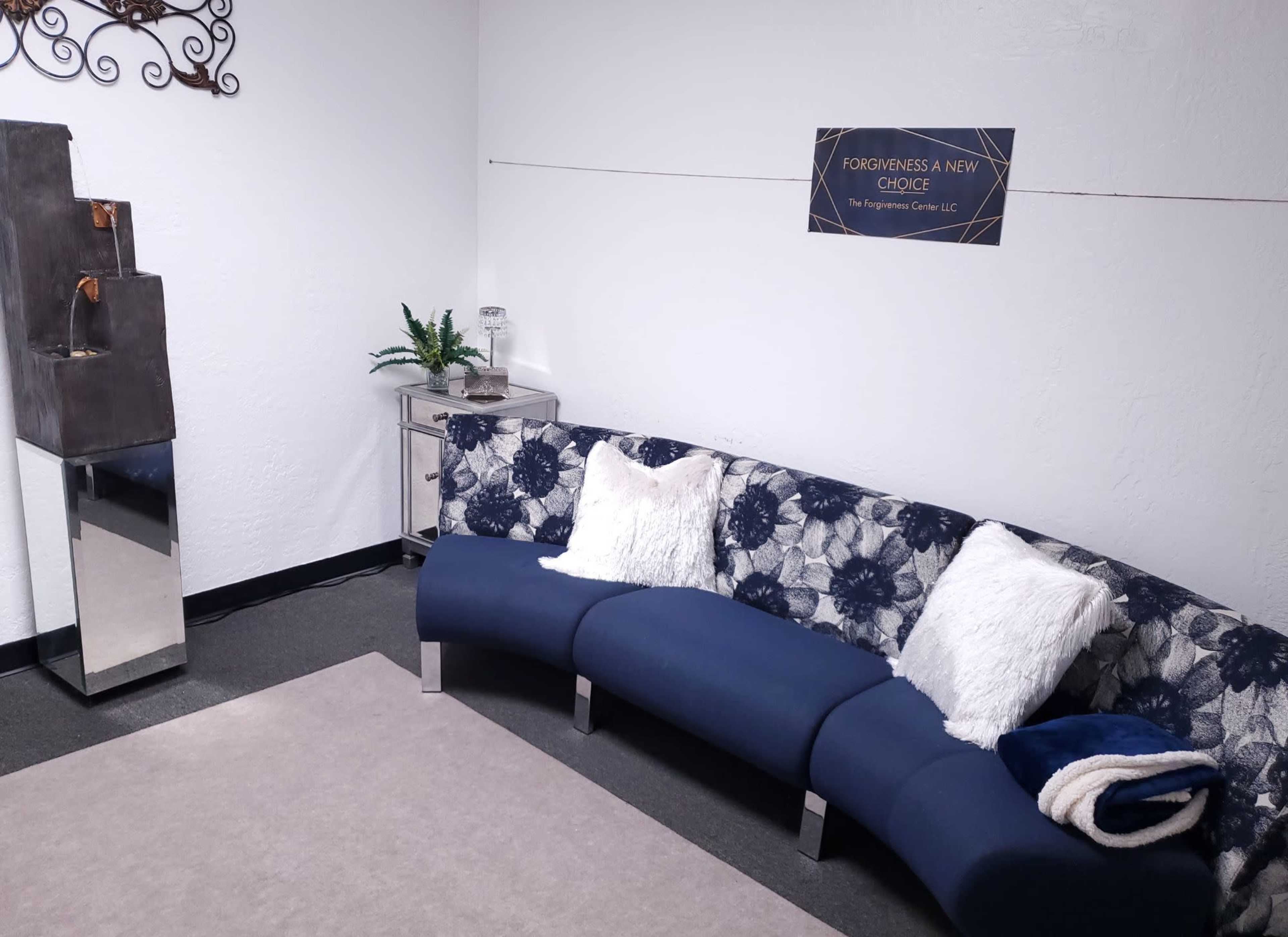 A modern sitting area with a curved blue couch, decorative pillows, a small table, and a sign on the wall that reads "FORGIVENESS A NEW CHOICE."