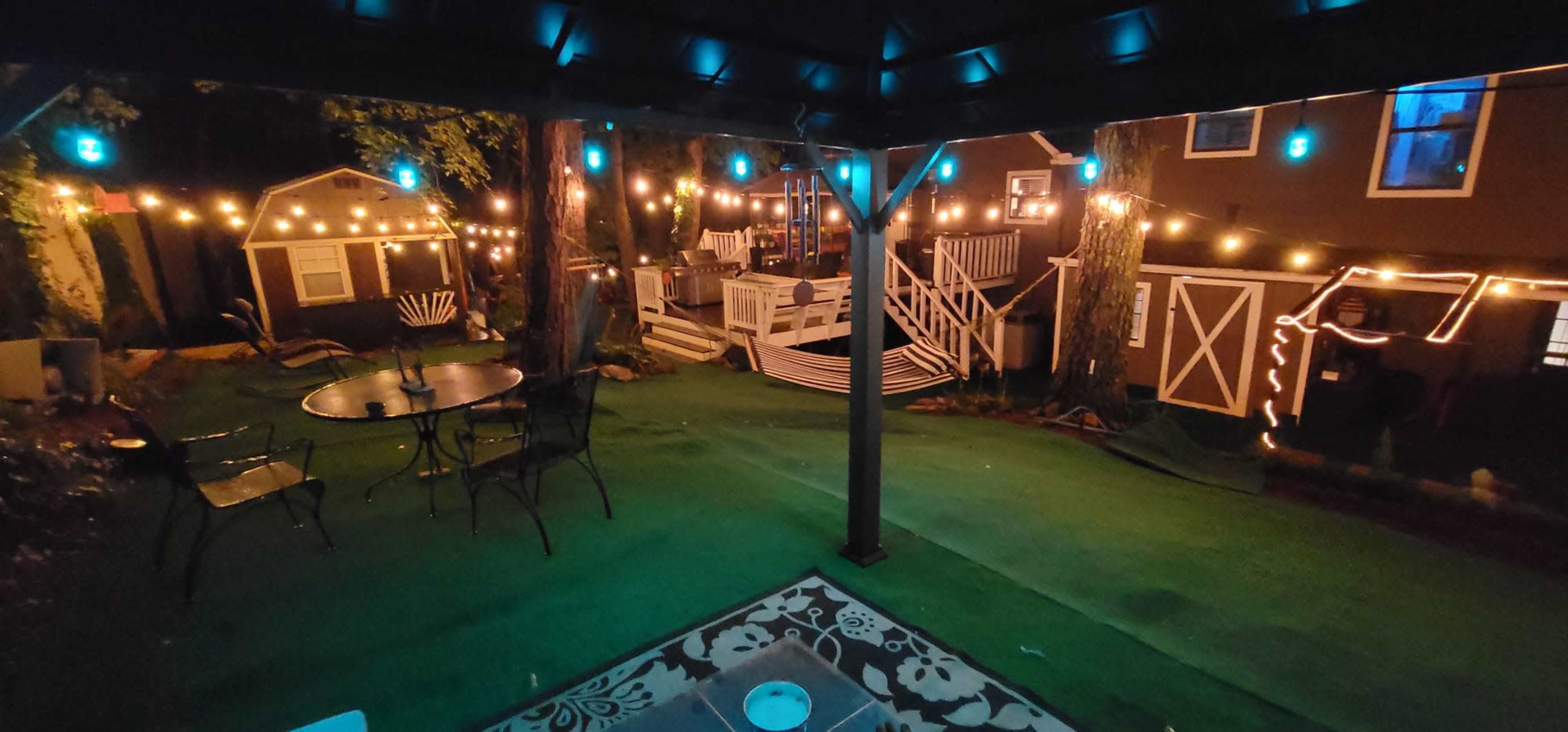 The scene shows a backyard decorated with string lights, featuring a patio area with a table and chairs, along with several small buildings visible in the background.