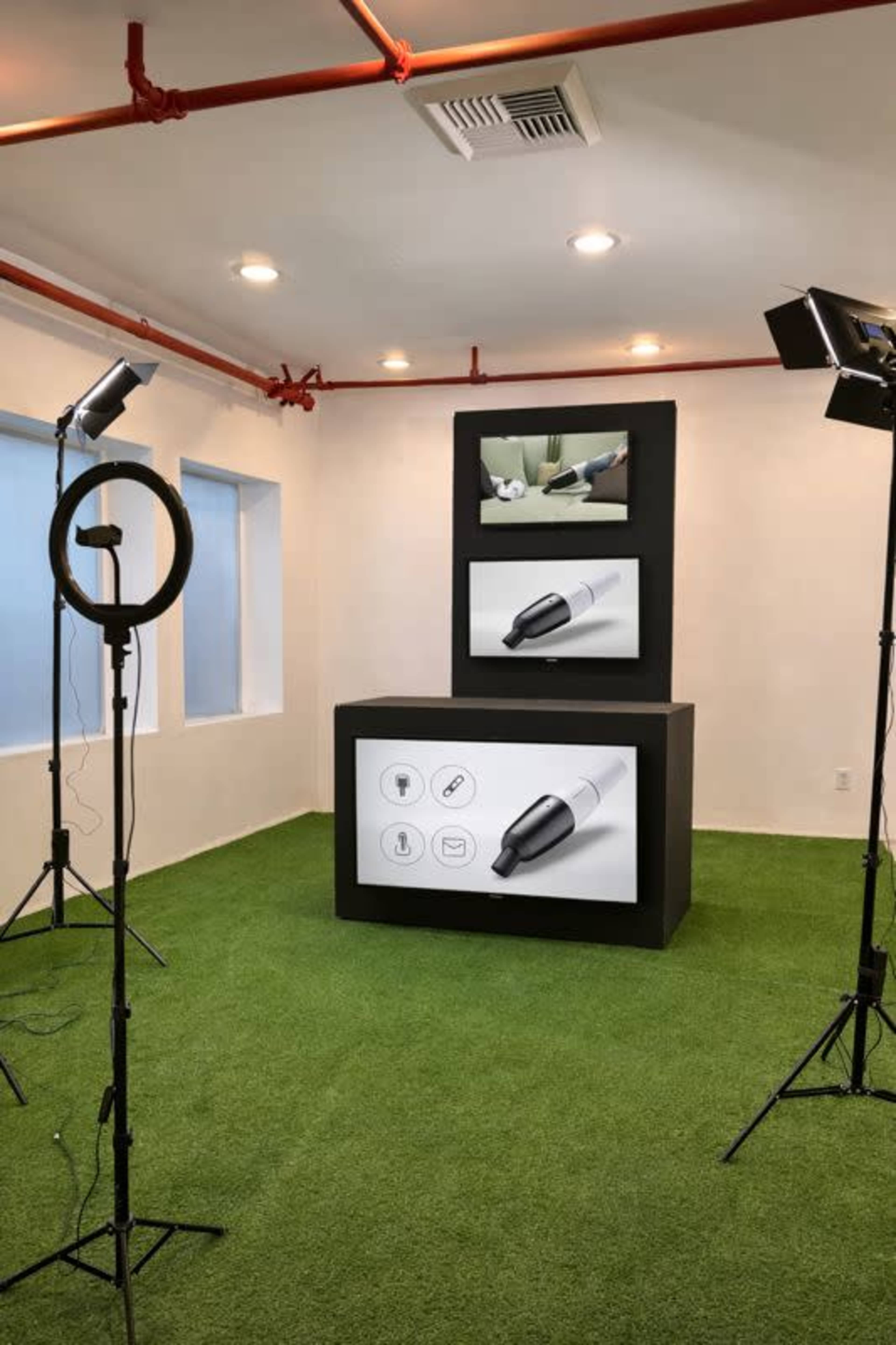 The image shows a modern exhibition space with a grassy floor, featuring a central display with screens and illuminated by two tripods with lights.