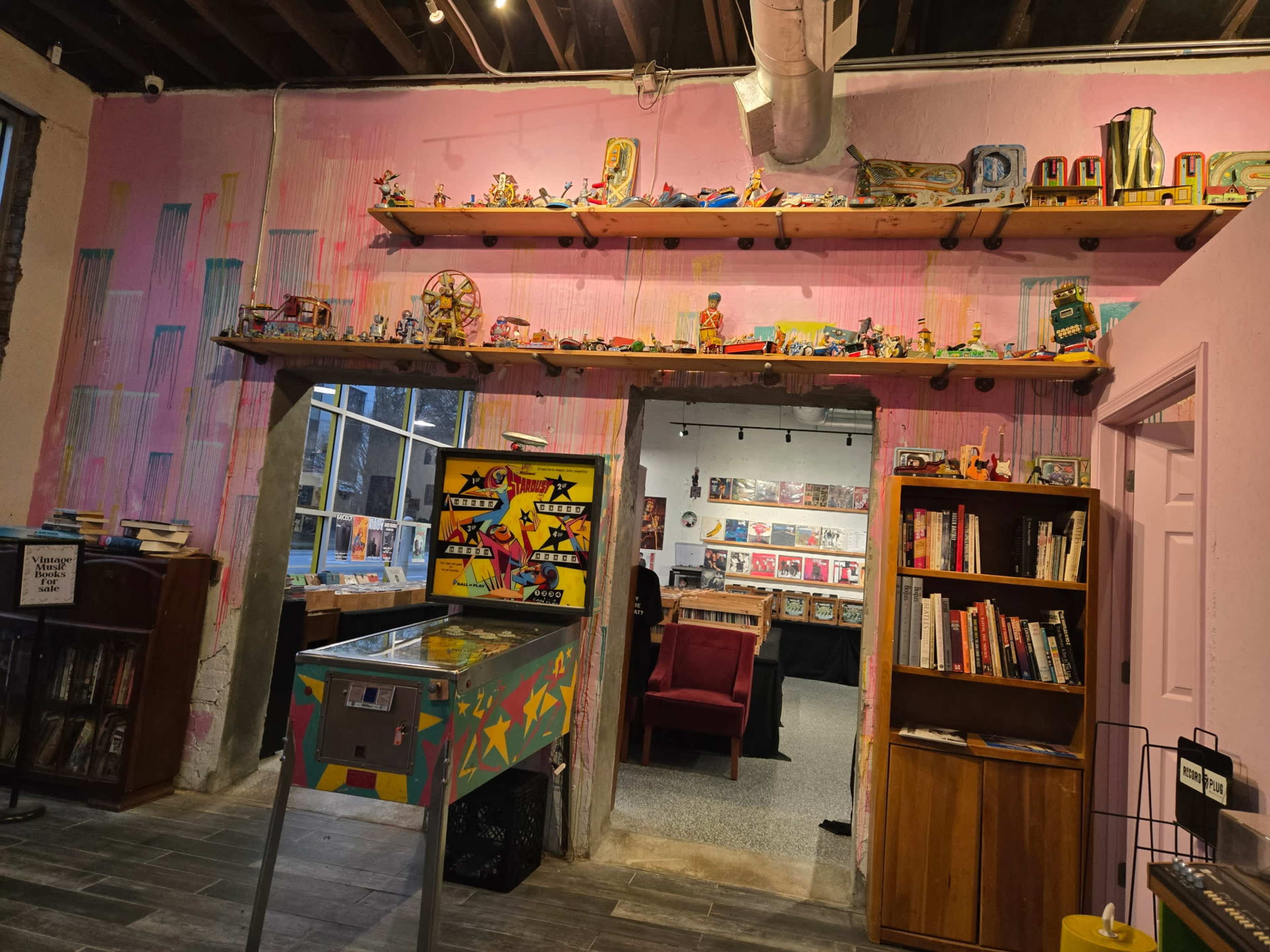 The image shows a brightly colored interior space with shelves displaying various collectibles, a pinball machine in the foreground, and a doorway leading to a room filled with books and seating.