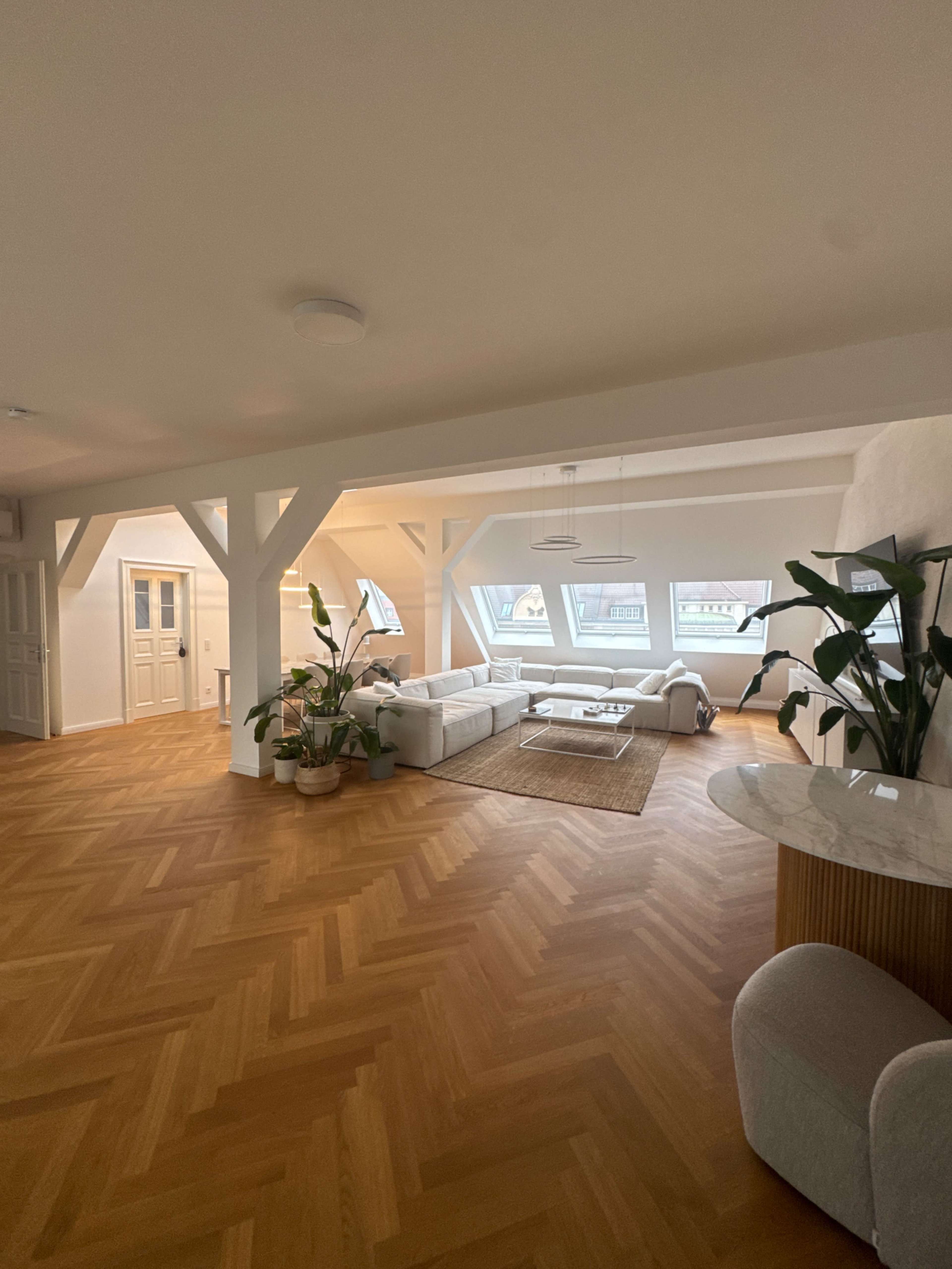The image shows a modern, spacious living room with large windows, light wooden flooring, and minimalist furniture, including a white sectional sofa and a round coffee table.