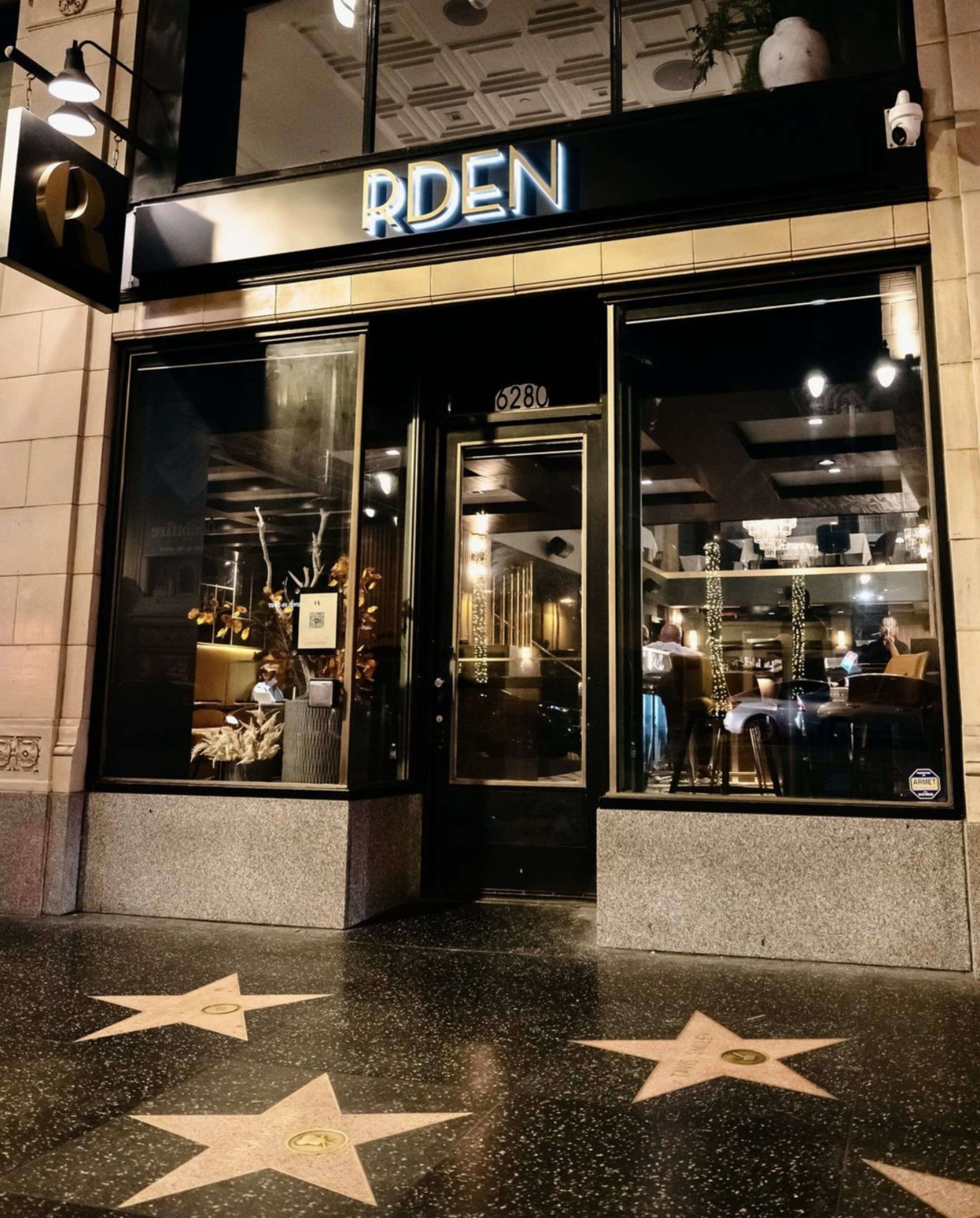 The exterior of a restaurant named "RDEN," featuring a glass entrance adorned with modern decor and located on a street with Hollywood Walk of Fame stars.