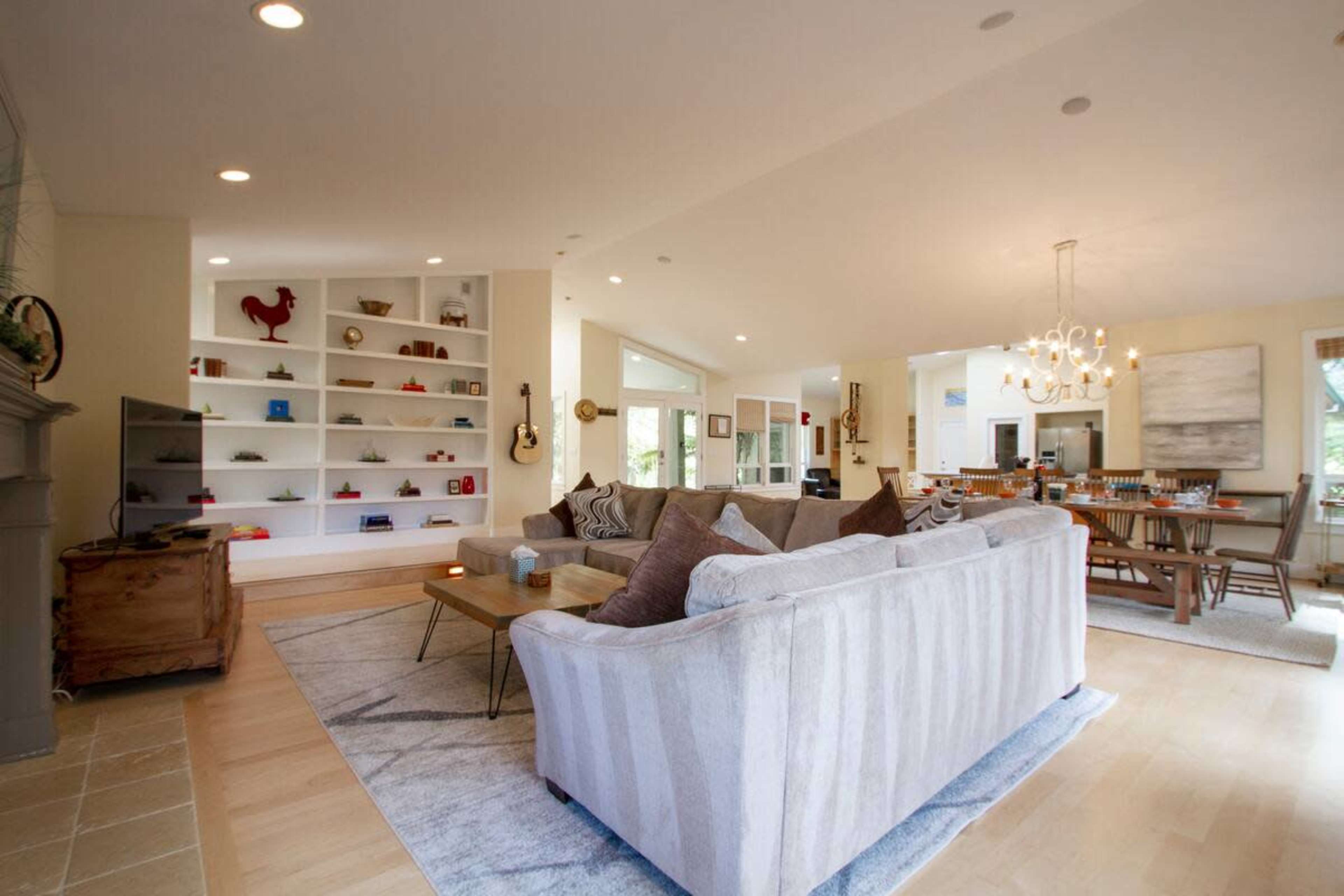 The image shows a spacious, modern living area featuring a light-colored sofa, a wall of decorative bookshelves, a dining area, and large windows allowing natural light to fill the space.