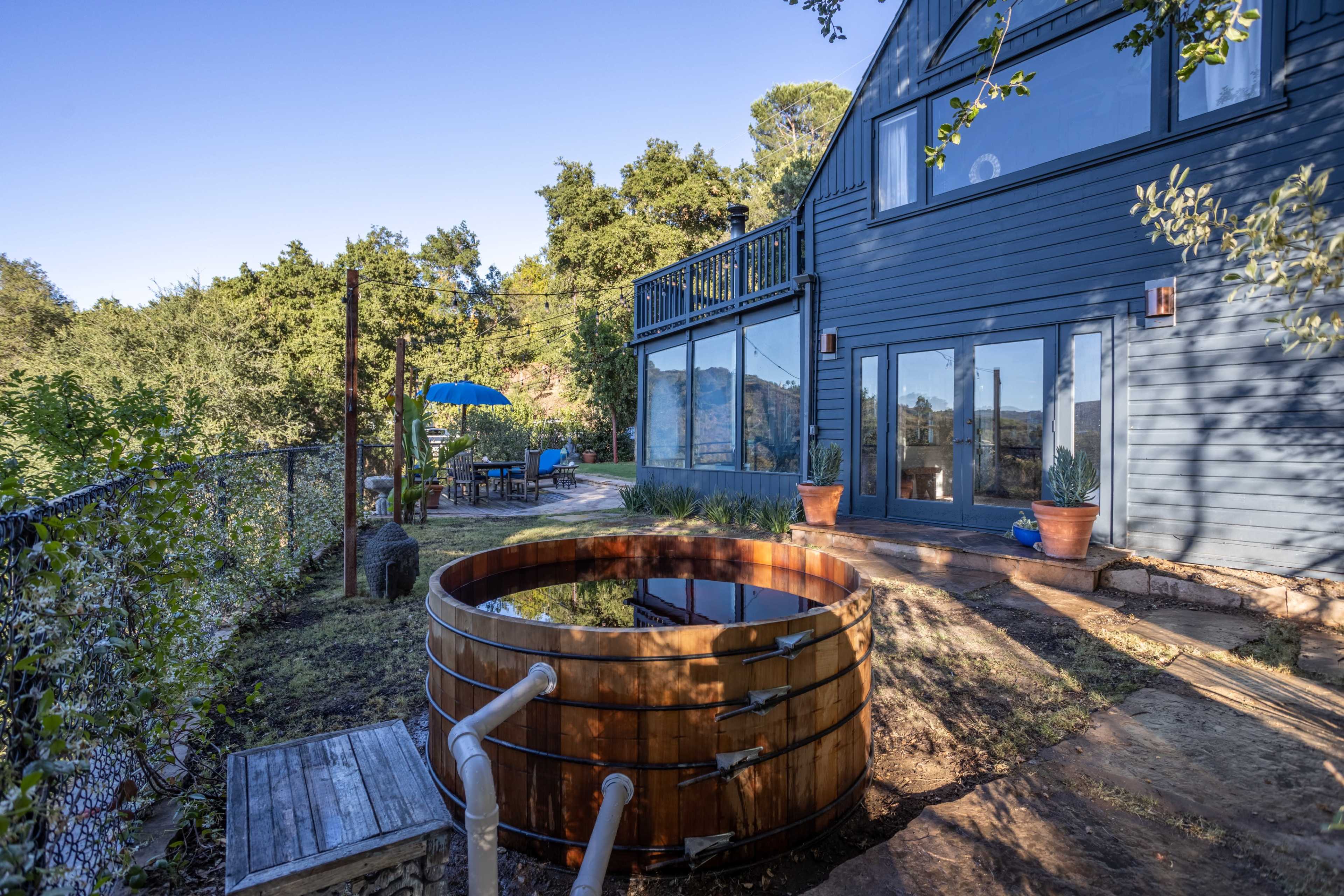 The image shows a wooden hot tub situated outside a blue house, surrounded by greenery and a stone path leading to the entrance.